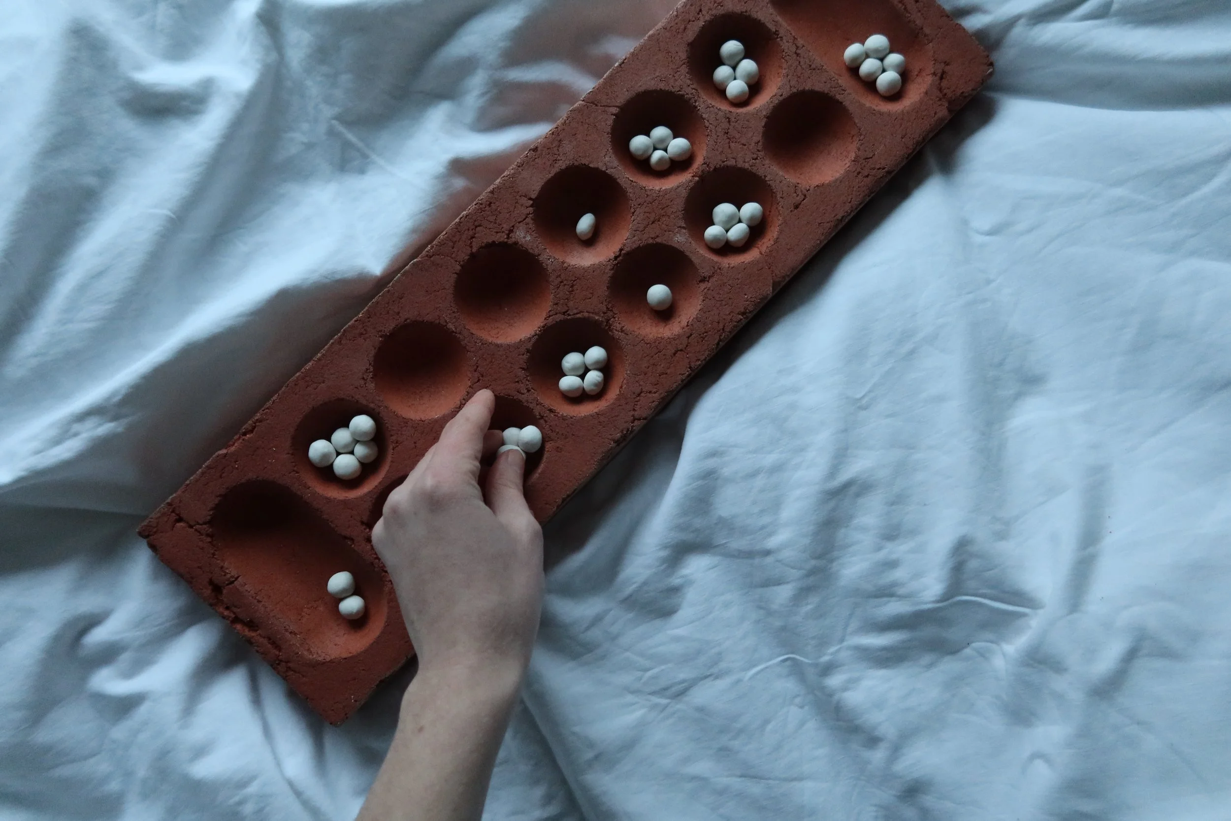 Hand legt Spielsteine aus Porzellan in die Mulden eines Mancala-Spielbrett aus rotem Ton.
