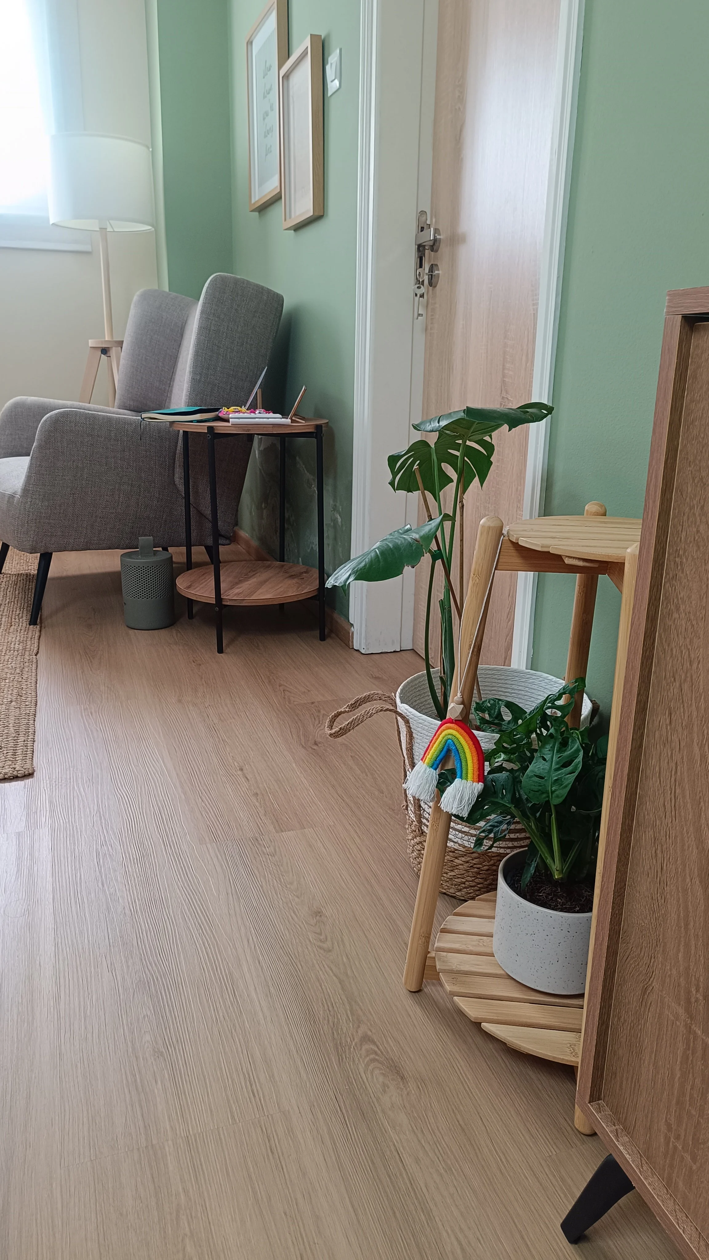 Living room corner with a gray armchair, a small black side table with art supplies, various framed pictures on a green wall, a tall white lamp, a potted monstera plant with a rainbow decoration, and a wooden shelf with a slatted top.