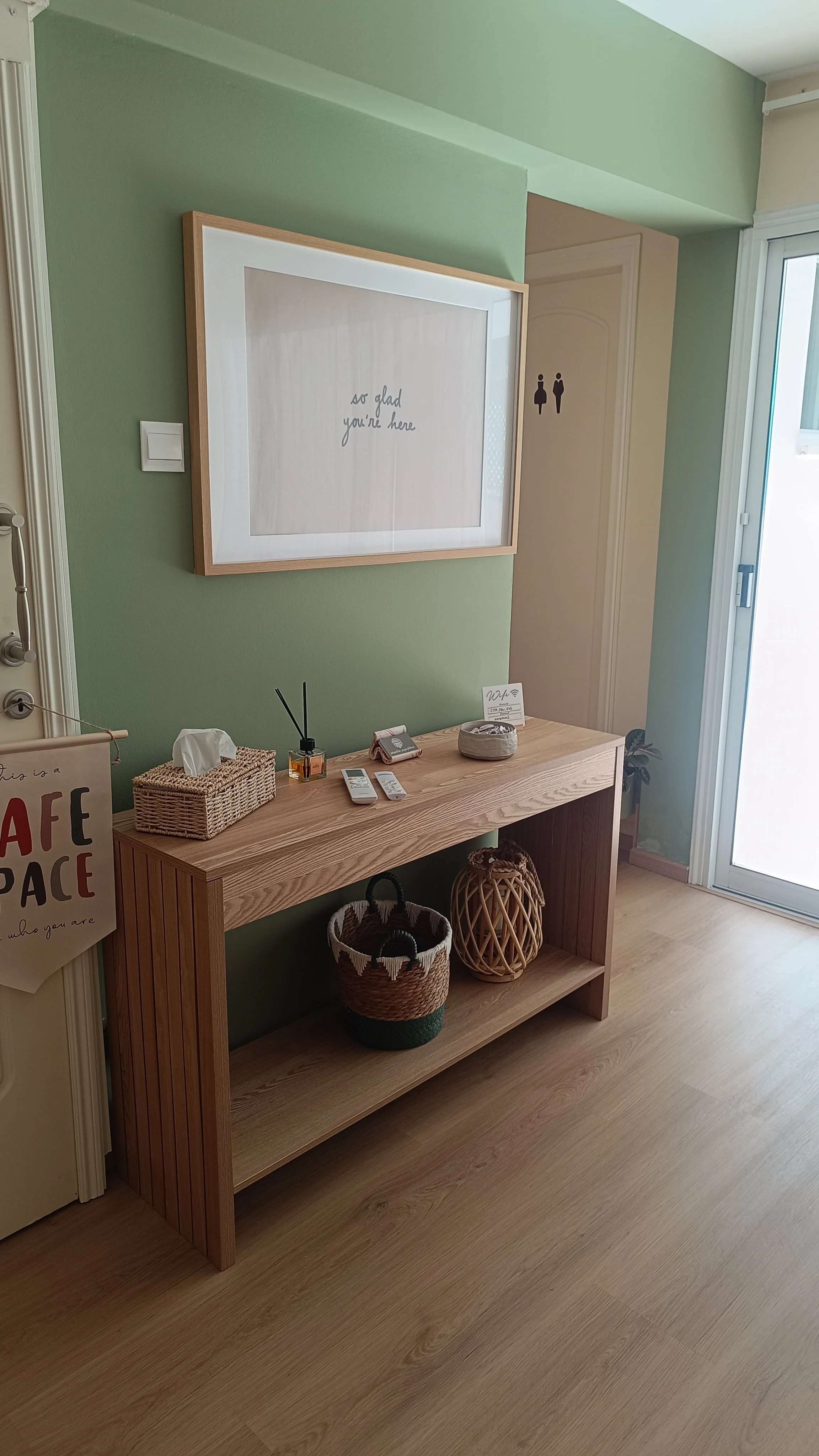 Wooden console table against a green wall with decorative baskets and small items on top, a framed art print with the text 'so glad you're here', a door with bathroom signs, and a sliding glass door to the right.