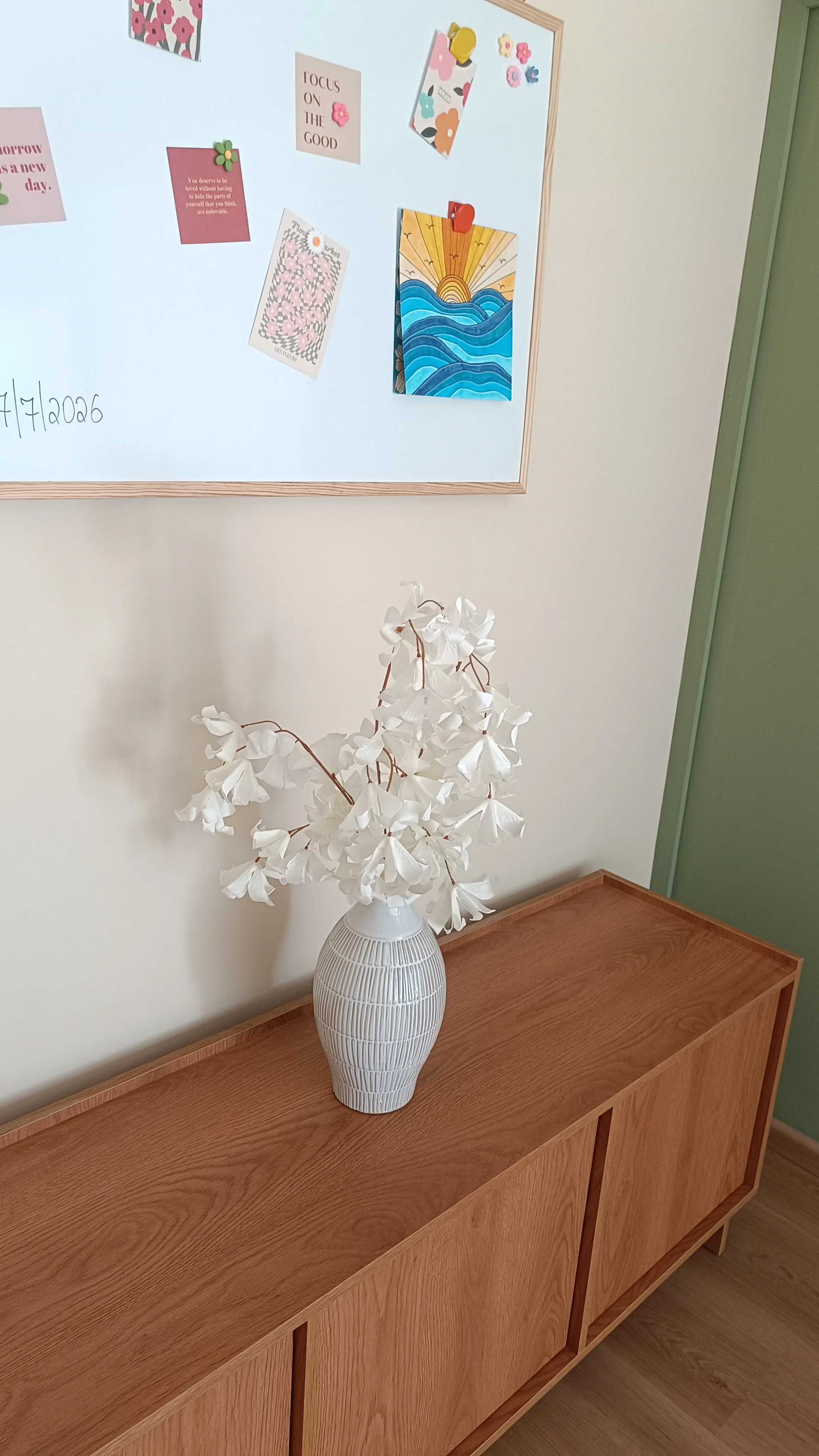 A white vase with a textured pattern holding white artificial flowers placed on a wooden sideboard in front of a white wall with a corkboard and decorative cards and pictures on it.