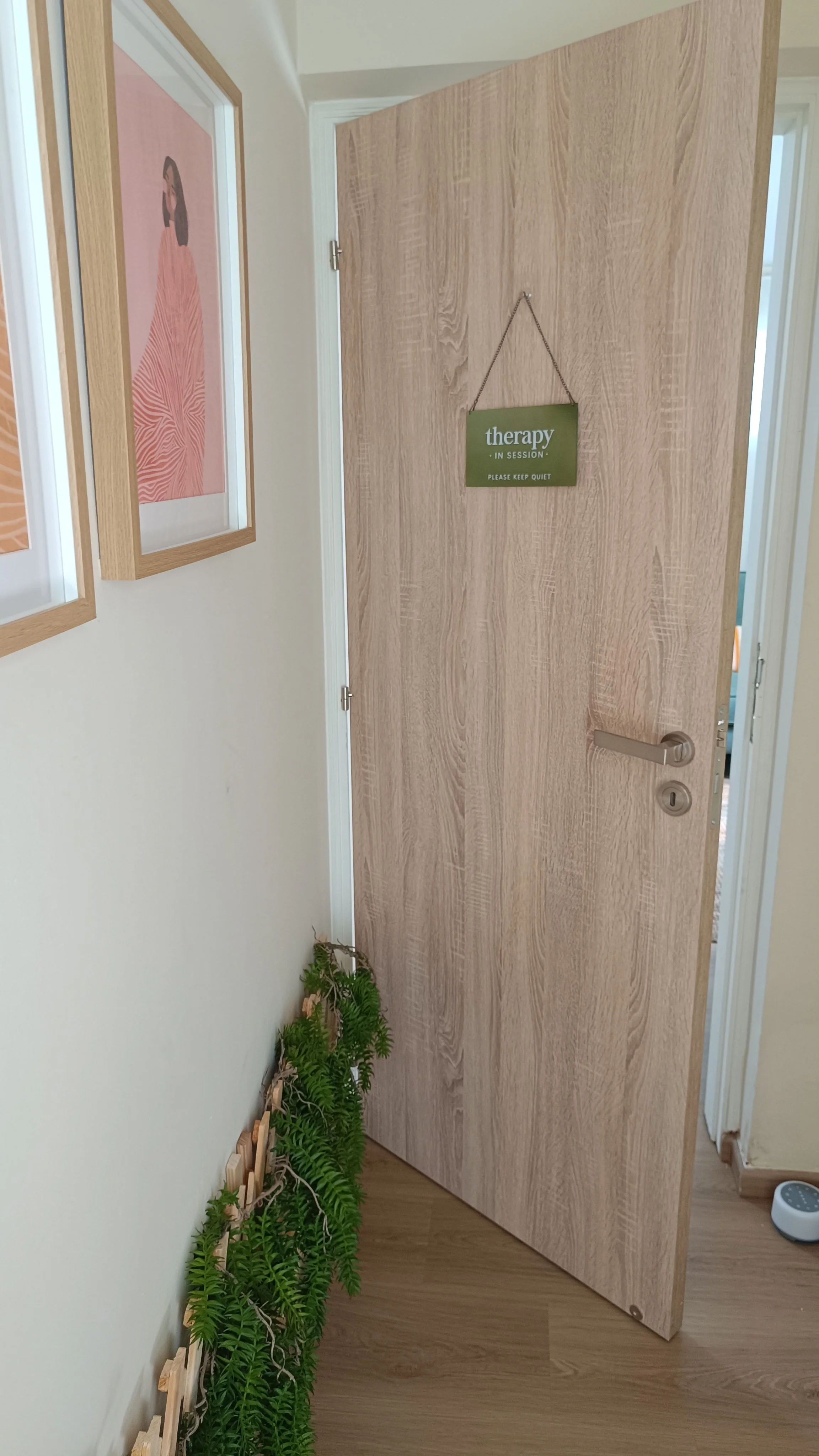 Open wooden door with a green sign that says "therapy in session, please keep quiet". Inside is a hallway with framed artwork on the wall and green plants lined up against the wall.