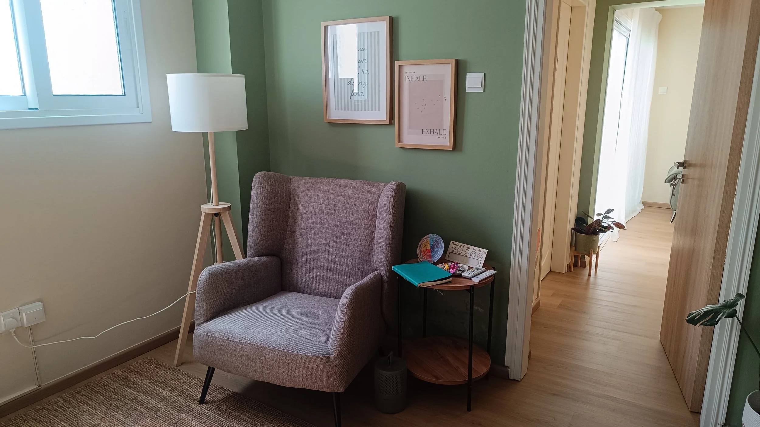 A cozy living room corner with a gray armchair, a wooden side table with a clipboard, remote controls, and a small plant, a tall white floor lamp, and framed artwork on a green wall, with a hallway visible in the background.