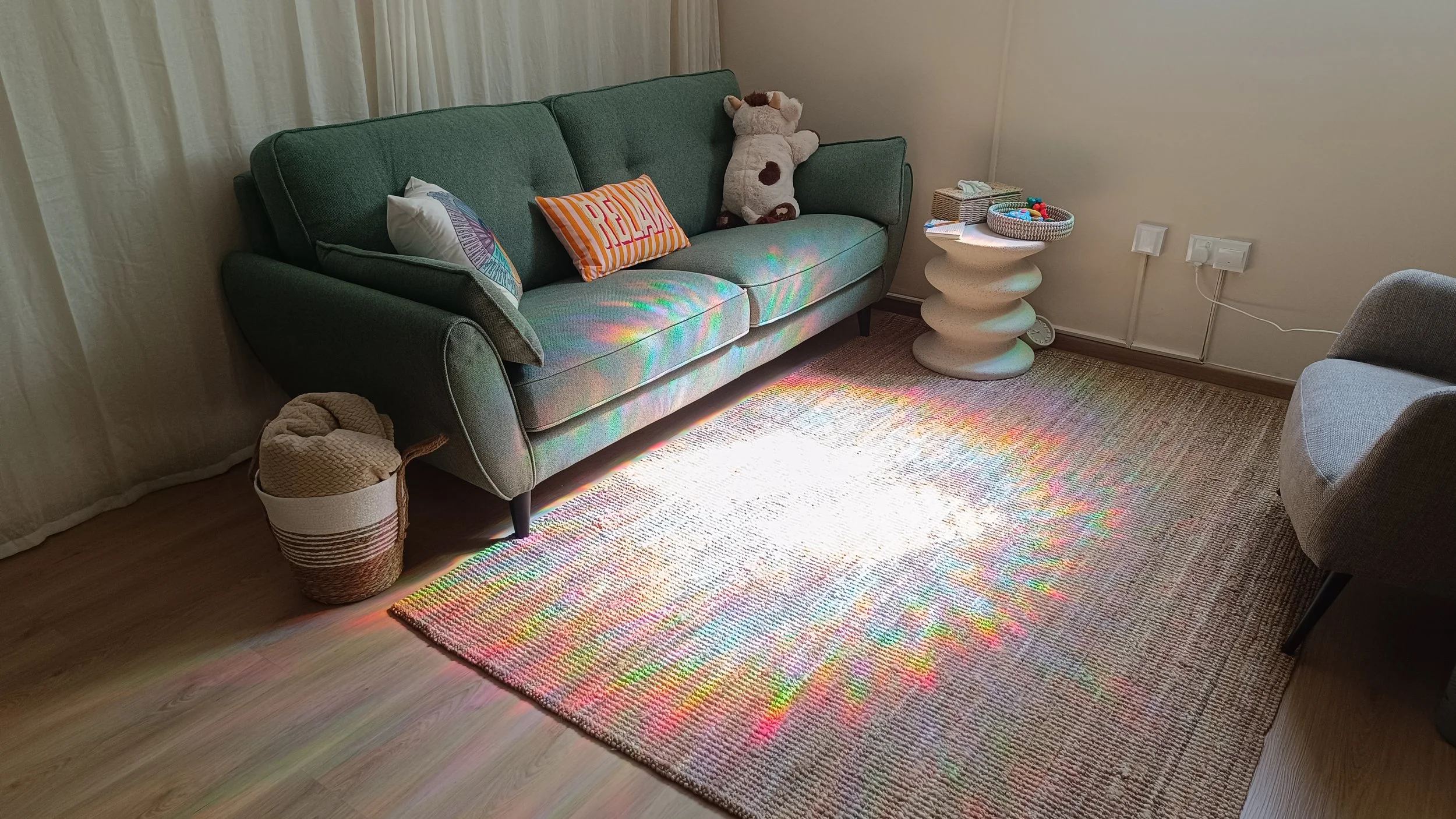 Living room with green sofa, colorful pillows, a plush cow toy, side table with basket toys, a woven basket, a gray armchair, a multicolored rug, and rainbow light reflections on the floor and sofa.