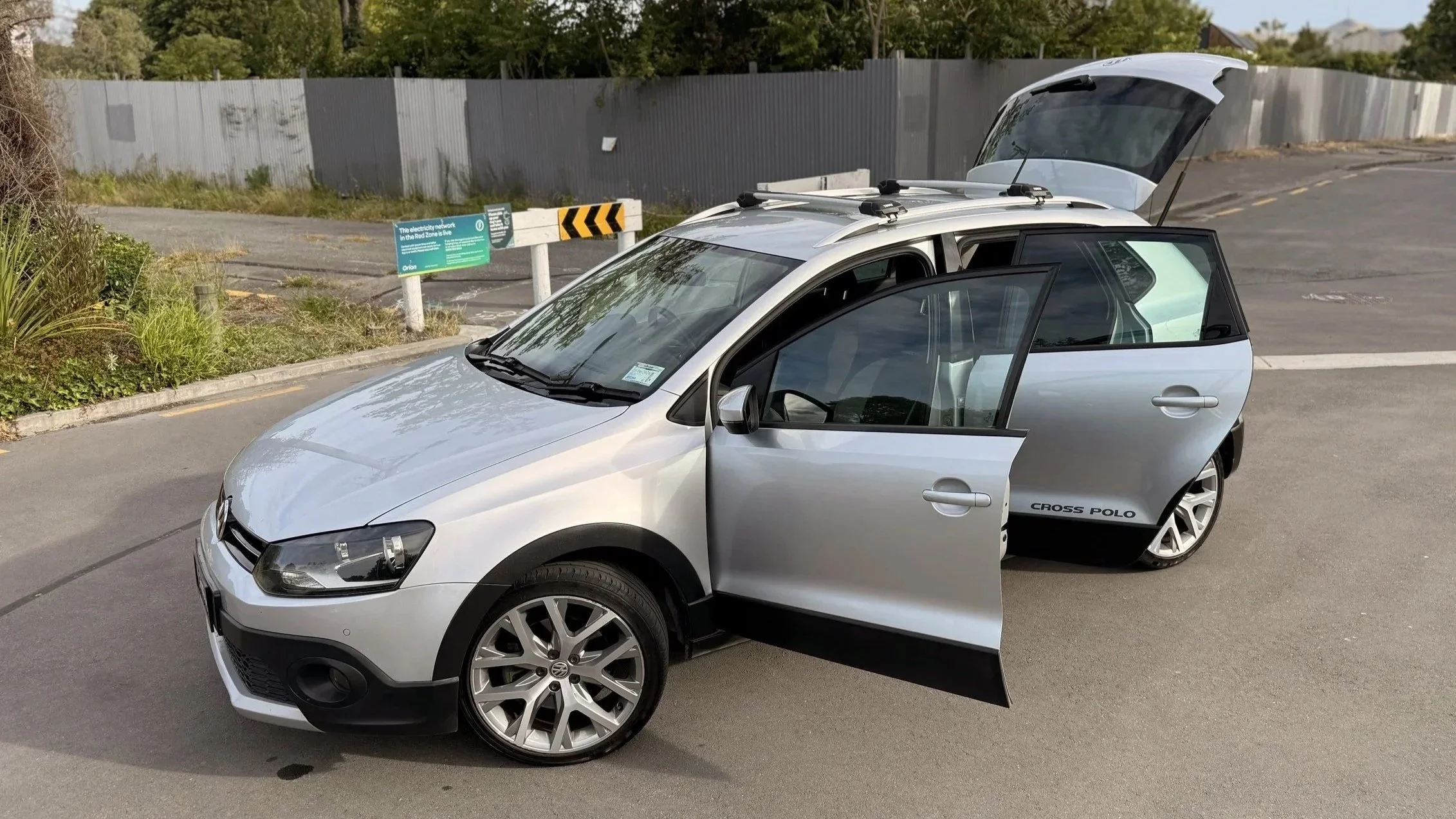 A silver Volkswagen Cross Polo with the driver's side door open and the rear hatch open, parked on a paved area near a sidewalk and green plants, with a gray fence and signage in the background.