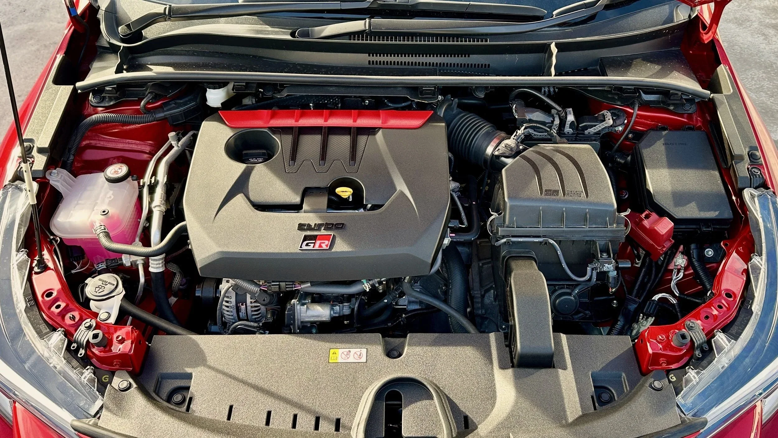 Engine bay of a red Toyota GR Corolla with a turbocharged inline-three engine, showing various mechanical components and fluid reservoirs.