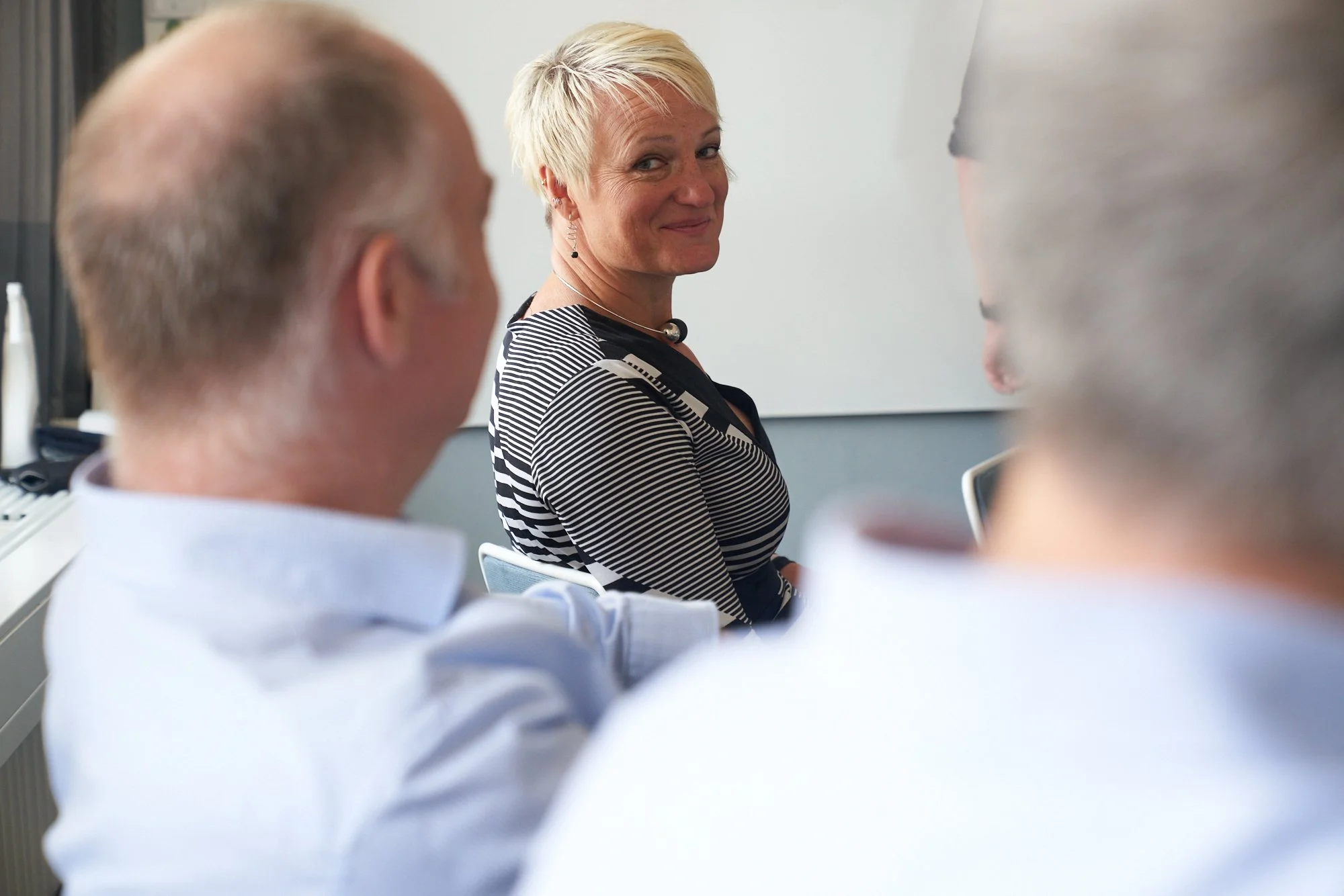 A woman with short blonde hair smiling in a meeting room surrounded by people.