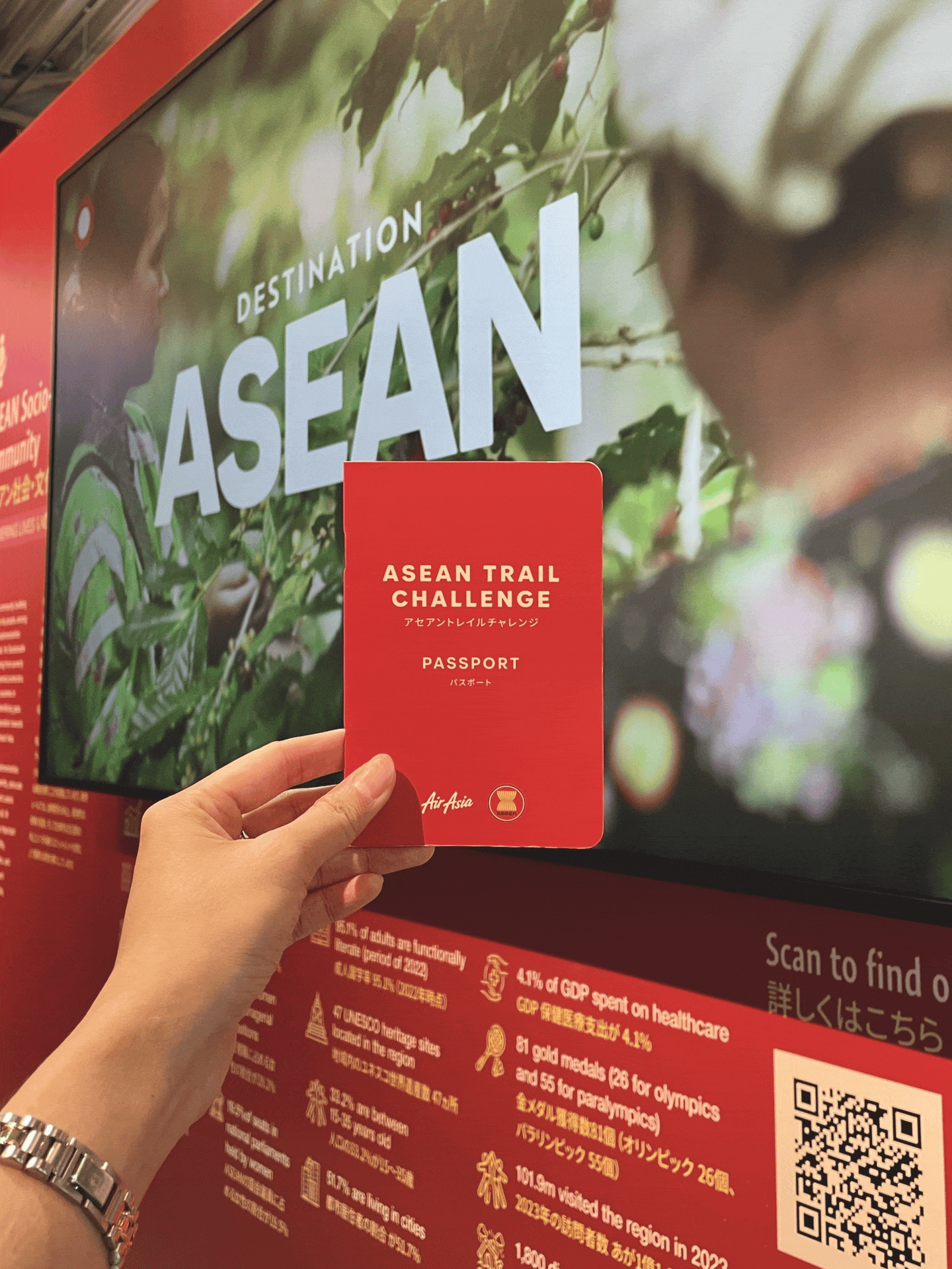 Person holding a red ASEAN Trail Challenge passport in front of an ASEAN-themed display at an exhibition.