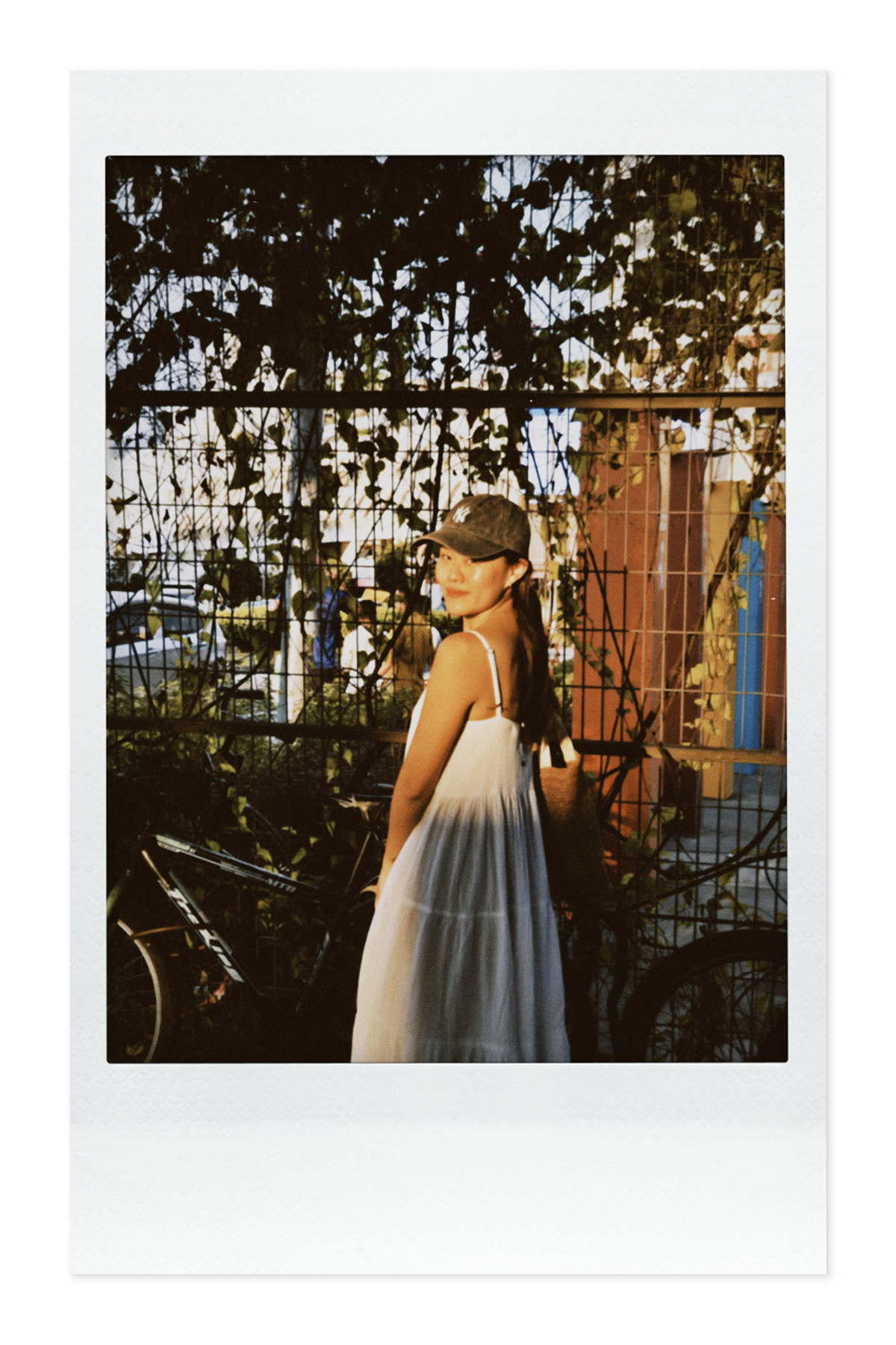 A woman wearing a white and dark-colored dress and a baseball cap, standing outdoors near a metal fence and trees, during sunset.