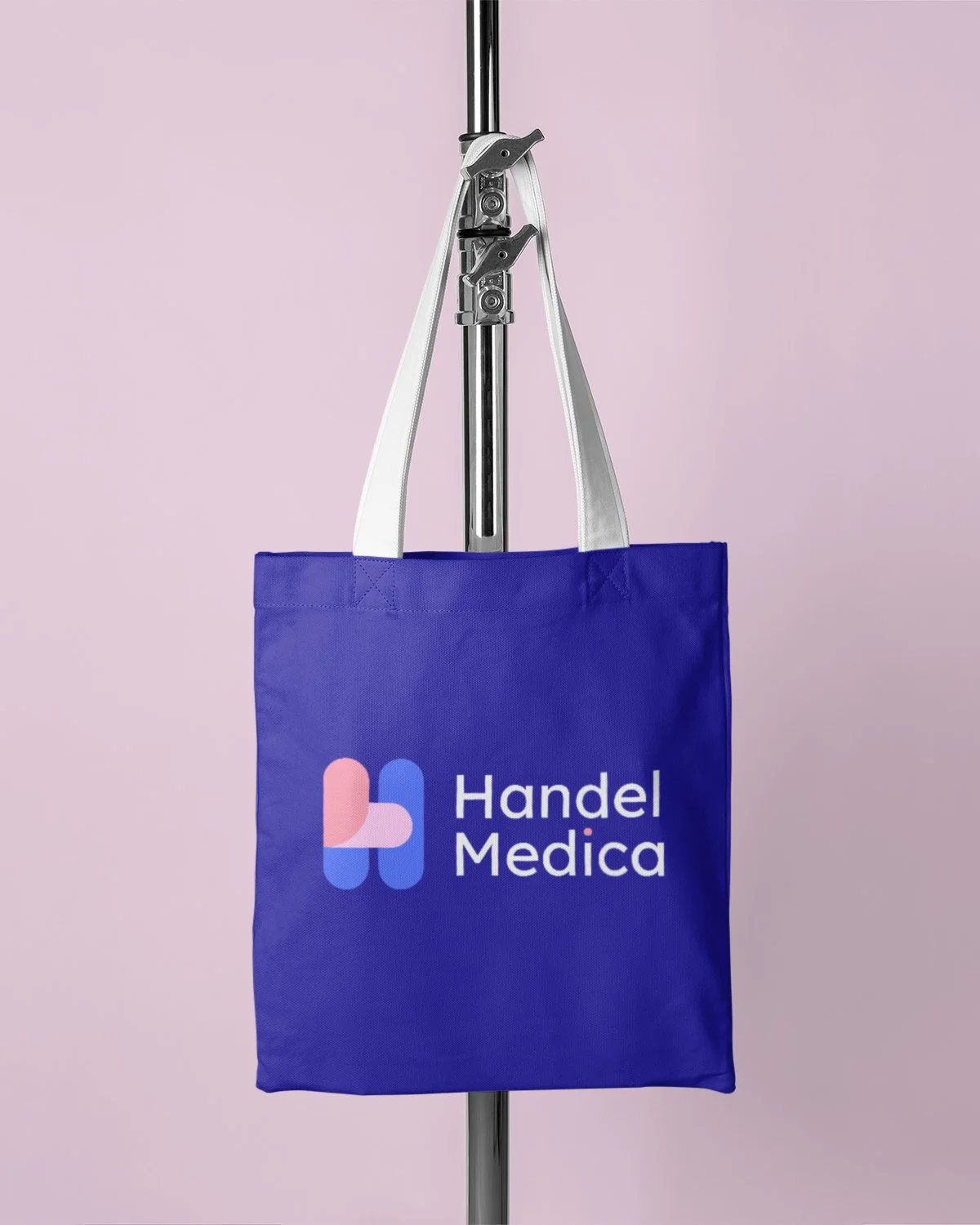 A blue tote bag hanging from a metal stand with a logo and text 'Handel Medica' on it, set against a light pink background.