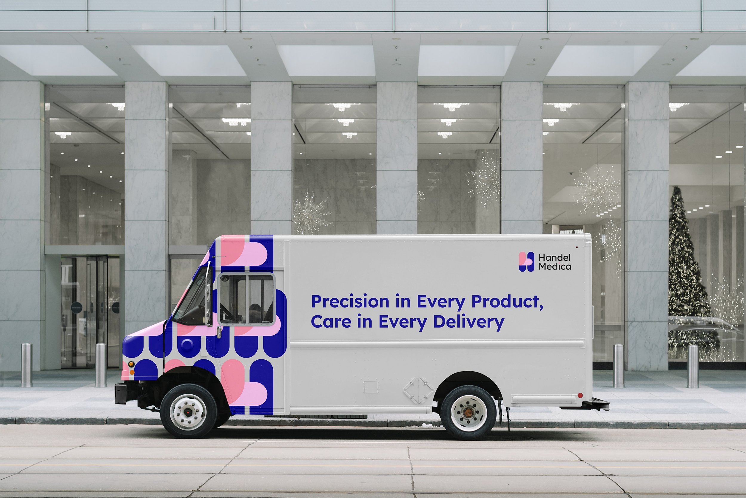 A delivery truck parked in front of a modern building with large glass windows, decorated with Christmas trees and holiday lights.