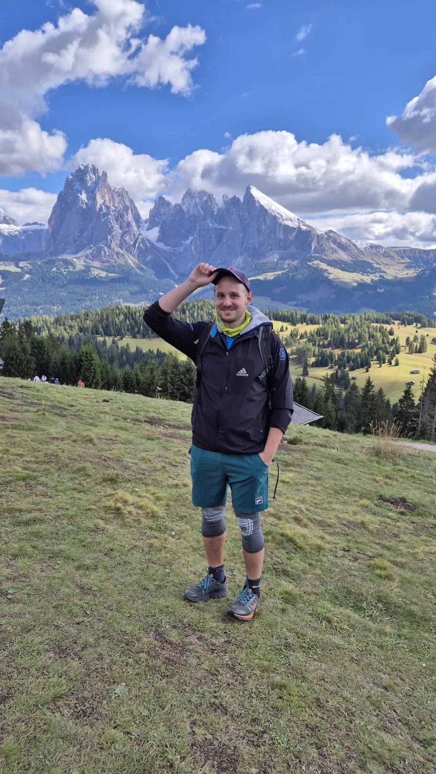 A man standing outdoors on grassy terrain, smiling and adjusting his cap, with a backdrop of towering snow-capped mountains, green forests, and a partly cloudy sky.