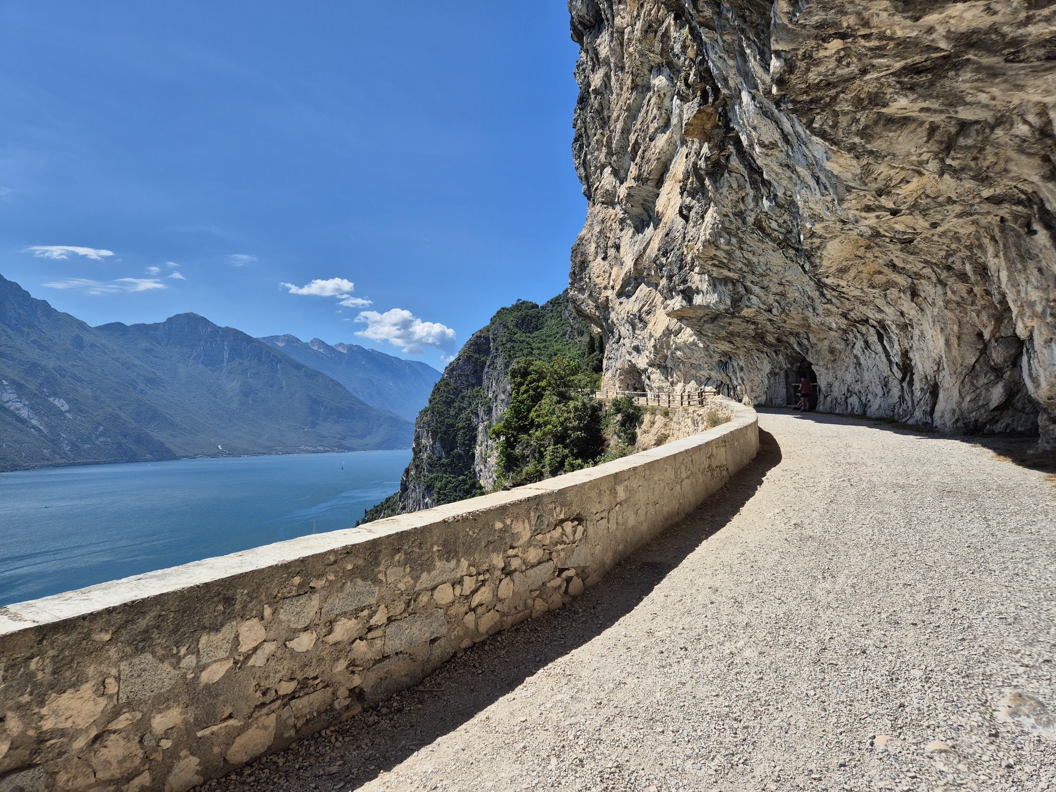 A gravel path along a rocky mountain ledge overlooking a river and distant mountains under a blue sky.