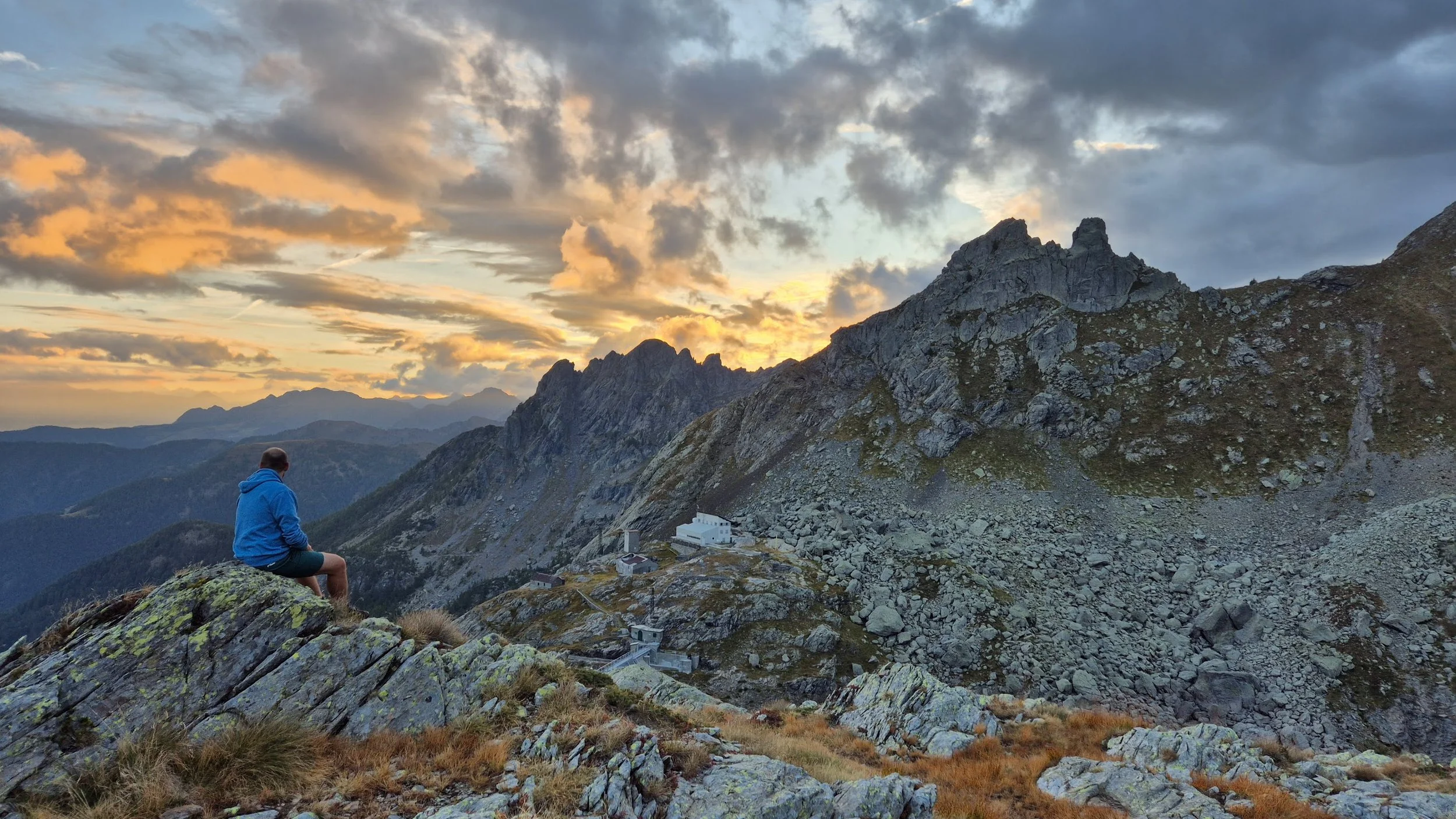 Un uomo seduto su una roccia guarda un paesaggio montano durante il tramonto, con nuvole colorate nel cielo e montagne al tramonto.
