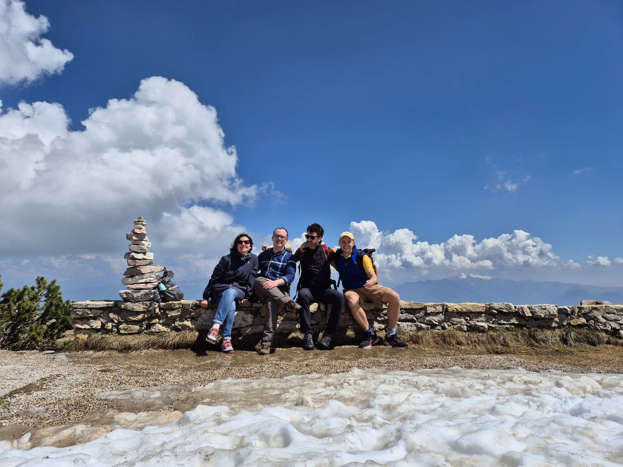 Four people sitting on a stone wall atop a mountain with a partly cloudy sky and distant mountain range in the background.