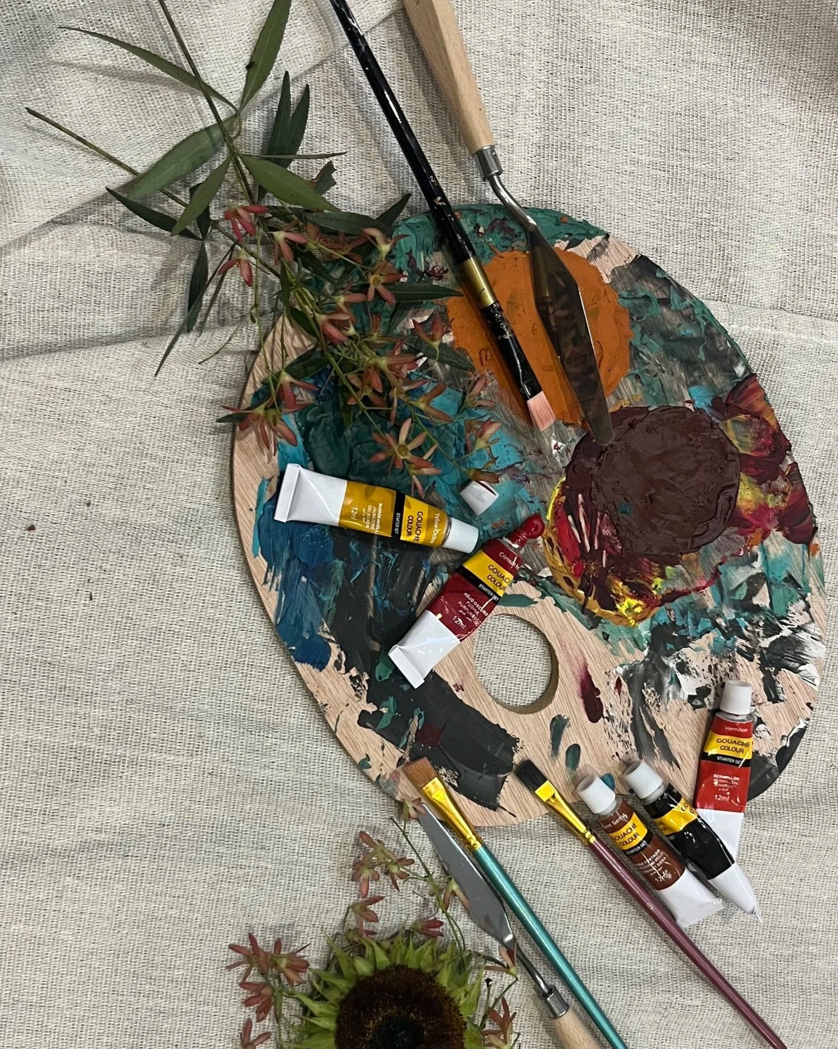 Painting supplies including a wooden palette with colorful paint, paintbrushes, tubes of gouache paint, and flowers on a fabric surface.
