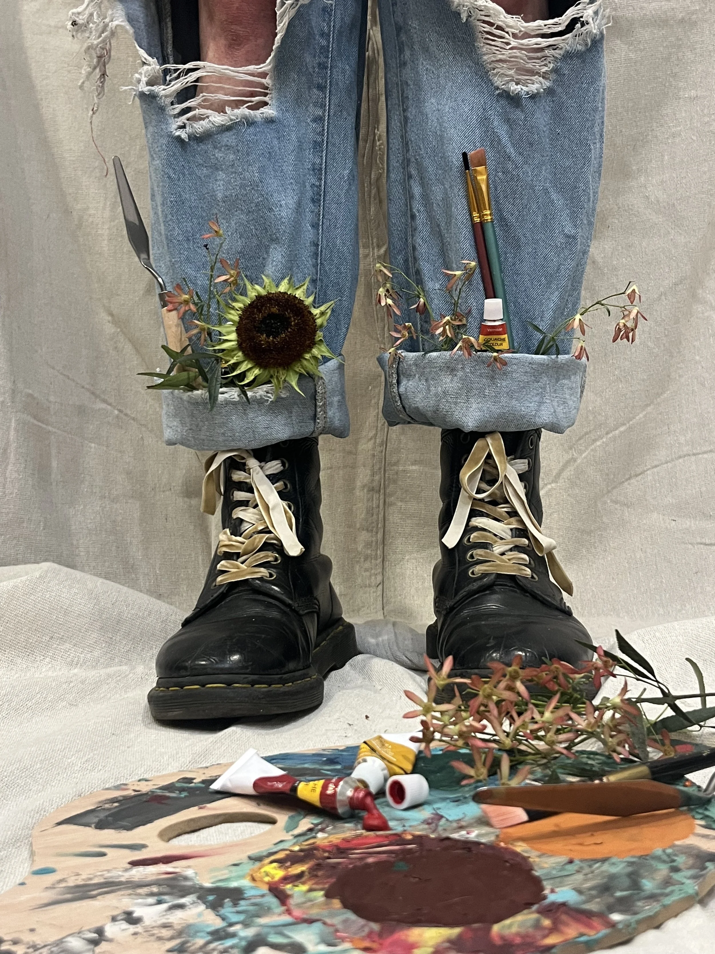 Close-up of a person's legs wearing ripped jeans and black boots, with small fabric pockets on the jeans containing a sunflower, plants, paintbrush, paint tube, and lighter, placed against a neutral background. An artist's palette with paint tubes, brushes, and a flower are in the foreground.