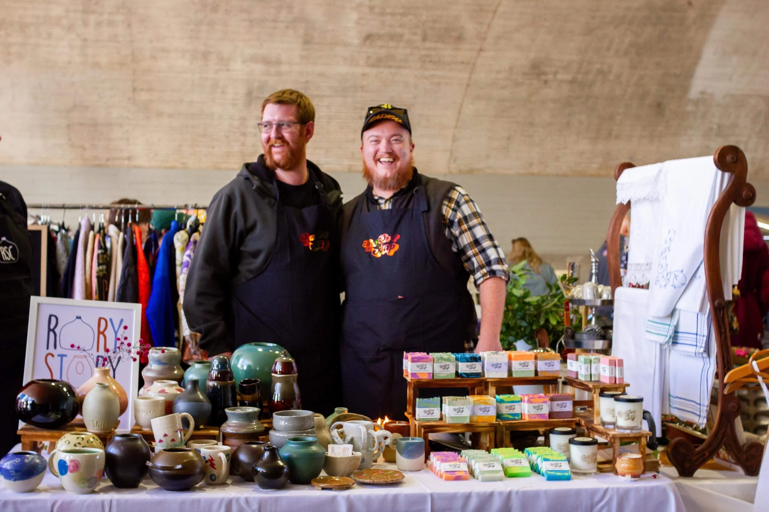 “Don’t Drop the Soap makers standing behind a market stall displaying handmade soap bars and artisan ceramics at a creative market.”