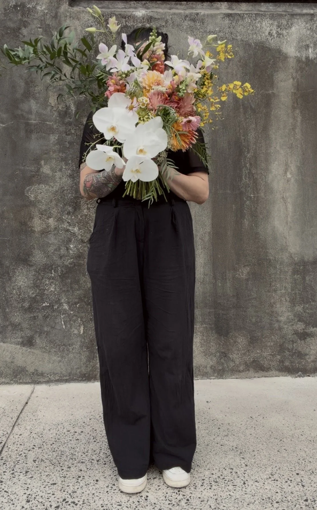 A person holding a large bouquet of flowers that covers their face, standing against a concrete wall. The bouquet includes white orchids, pink, yellow, and orange flowers, with green leaves.