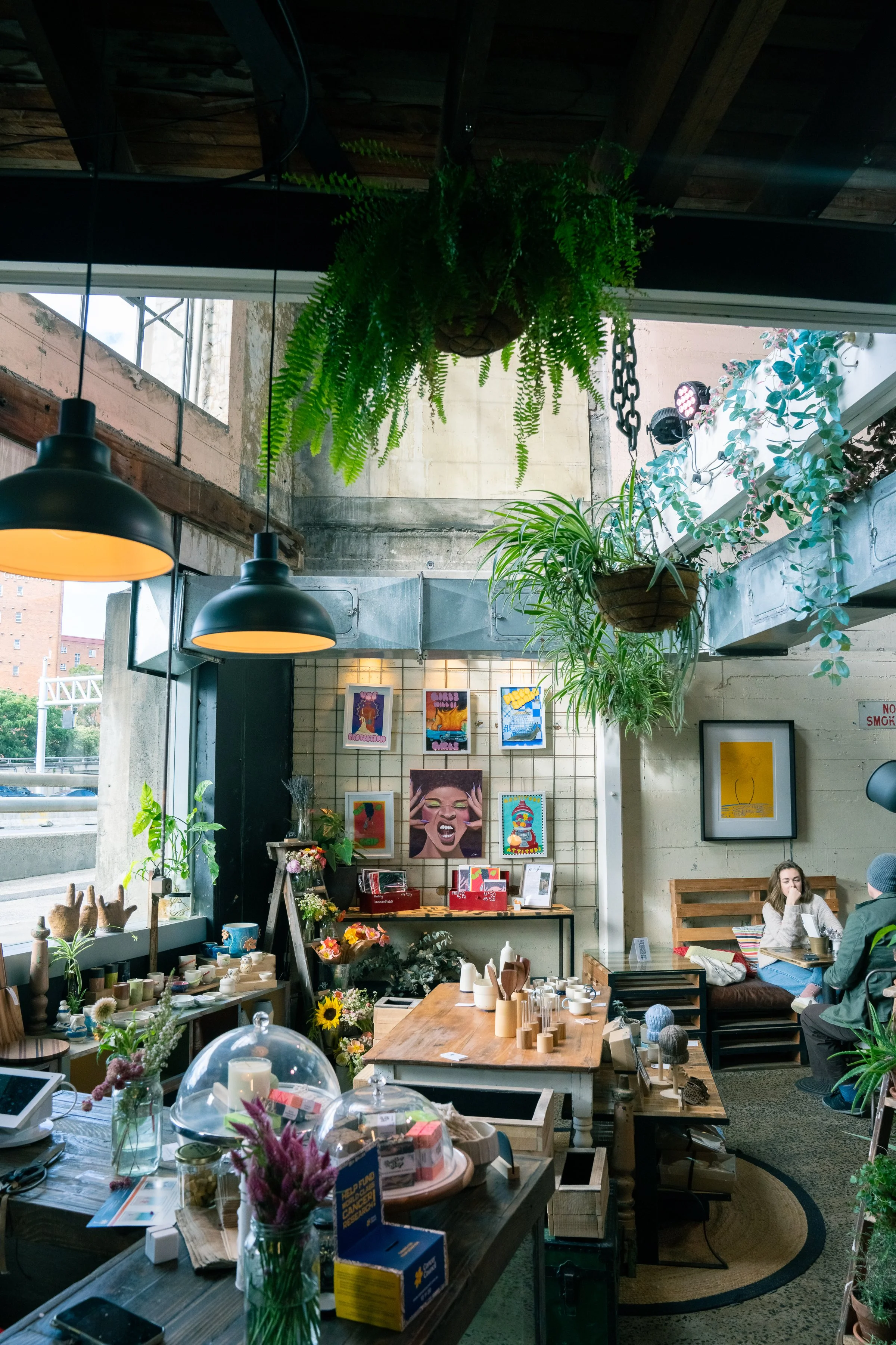Inside a cozy shop with hanging plants, framed artwork, and various items on display, with two people sitting at a table in the corner.