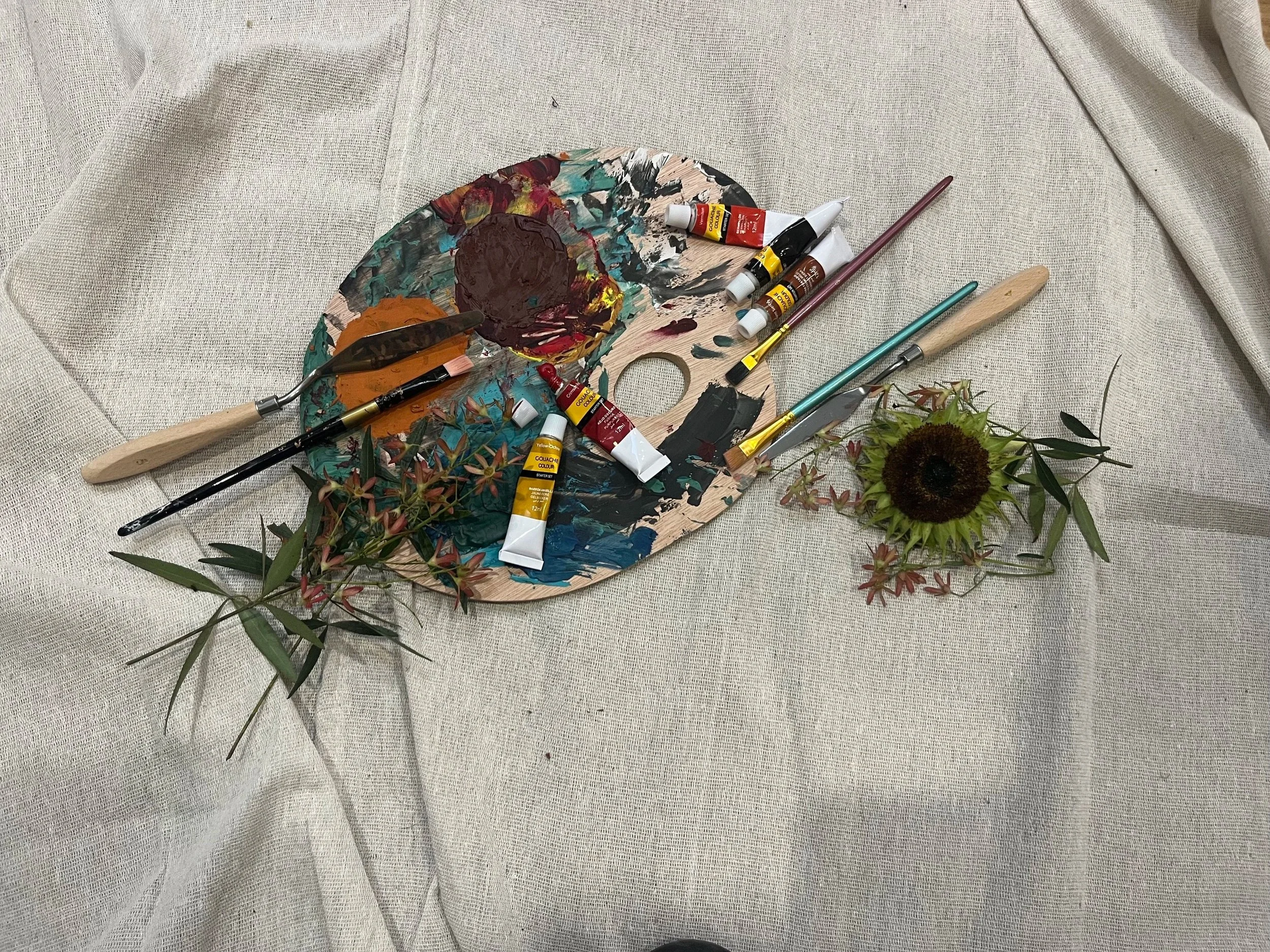 Art supplies on a beige fabric surface, including a wooden palette with colored paint, brushes, and tubes of acrylic paint, with a sunflower and greenery nearby.