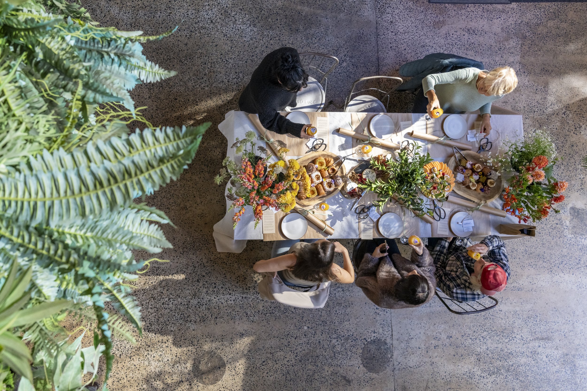 Overhead view of a dining table set with plates, napkins, and floral arrangements, with several people sitting and standing around, some holding drinks, in a well-lit indoor space with a speckled floor.