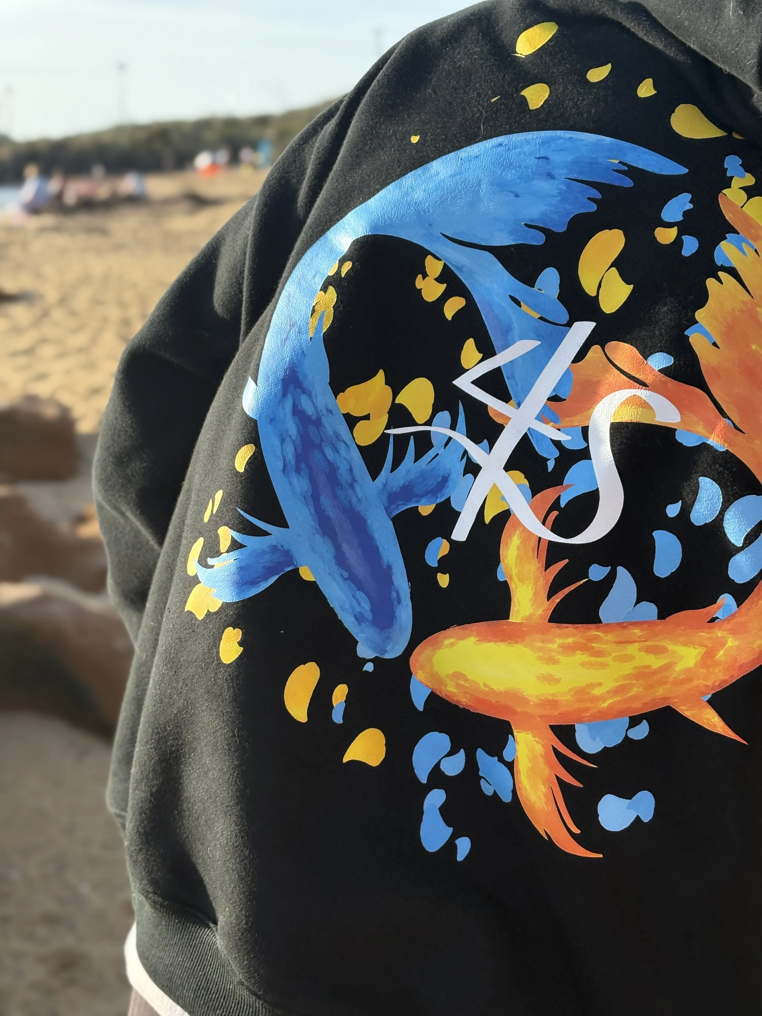 Close-up of a jacket with a colorful graphic of a fish on the back, featuring a blue and orange fish swimming in a black background with blue and yellow splashes, in a beach setting with sand and people in the background.