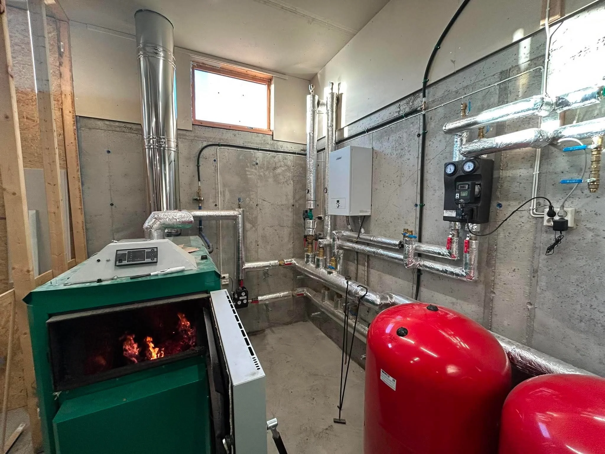 Et rom med isolerte rør, elektriske komponenter, og en peis som har ild, metallrør og redskaper på veggen, samt store røde tanker og en hvit kartong. Vindu øverst tillater lys inn i rommet.