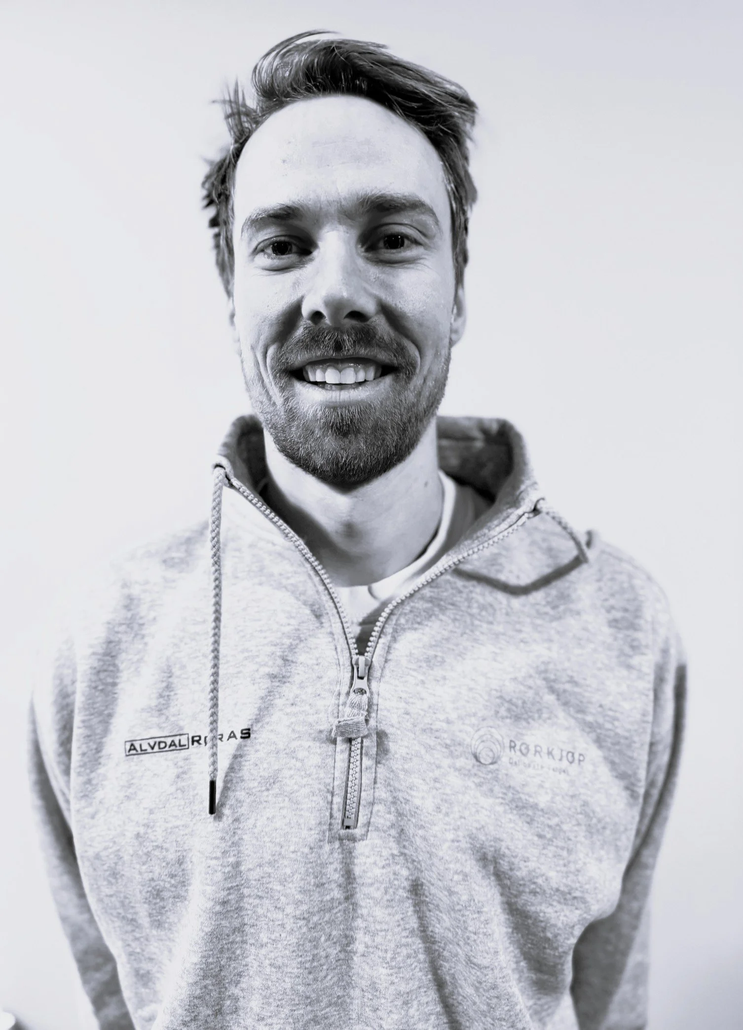 Erlend Lyster Ryen. En mann med lettere hår og skjegg, smilende og kledd i en lys grå hettegensrer med glidelås, står mot en enkel, lys bakgrunn.