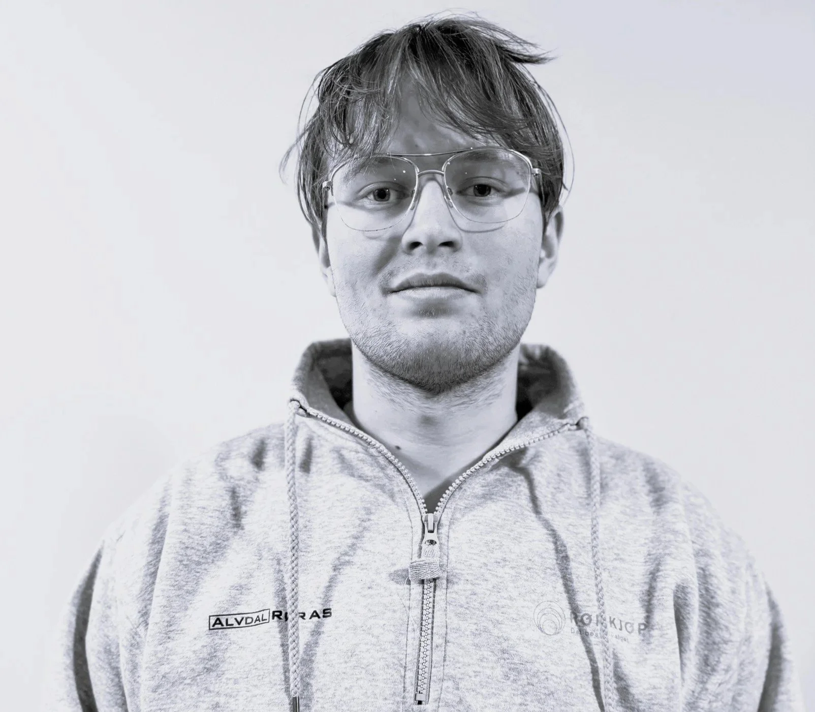 Konrad Streitlien. Sort-hvitt portrett av en ung mann med brunt, litt bustete hår, iført store, runde brillene og en lys grå hettegenser med glidelås. Han står foran en enkel, lys bakgrunn.