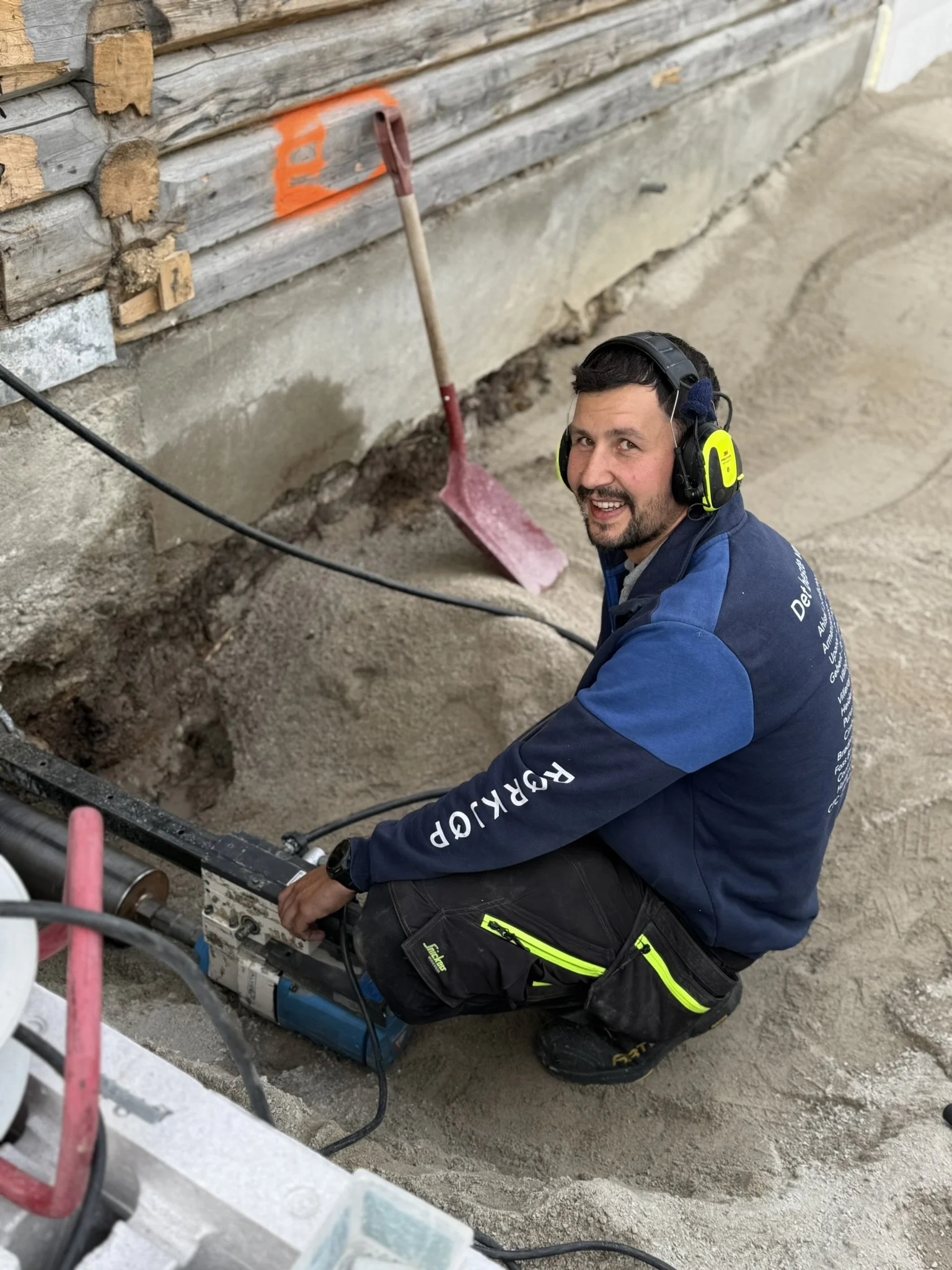 En mann som arbeider med å kjerneborre i en eldre bygning utstyrt med hørselsvern, foran en grov betongvegg og en utgravning.