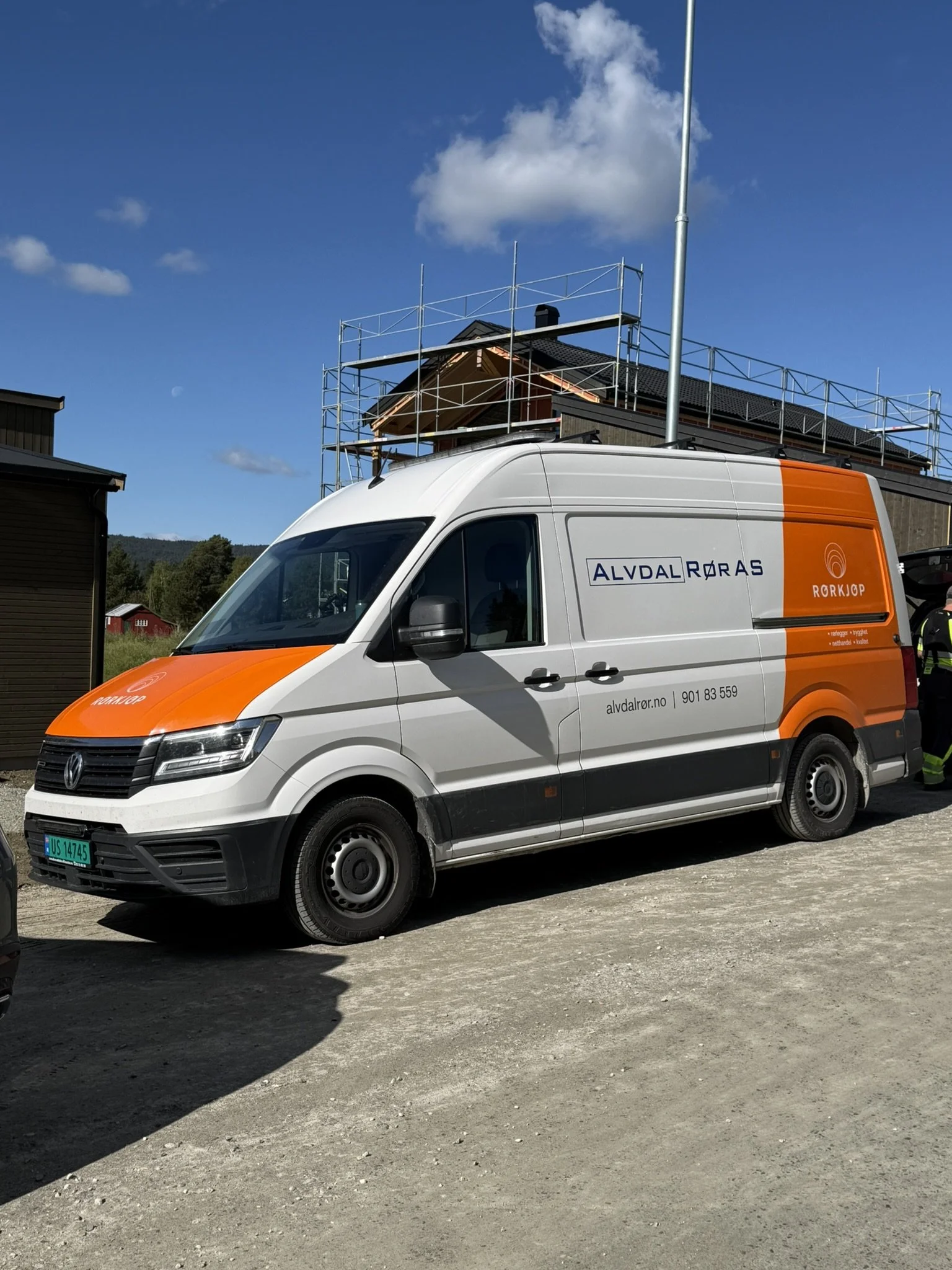 En hvit og oransje varebil med påskriften 'ALVDAL RØR AS' står parkert på en jordvei. Det er byggearbeid på en bolig i bakgrunnen, omgitt av atmosfærisk klar himmel med noen skyer.