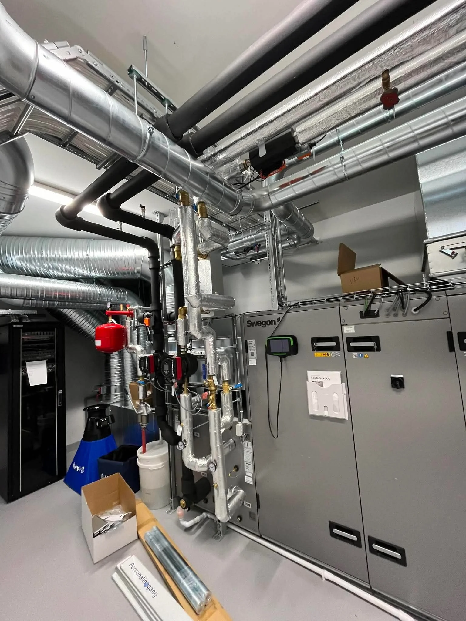 Interiør av teknisk rom med mange koblede metallrør, en elektrisk tavle, verktøy og utstyr på gulvet.