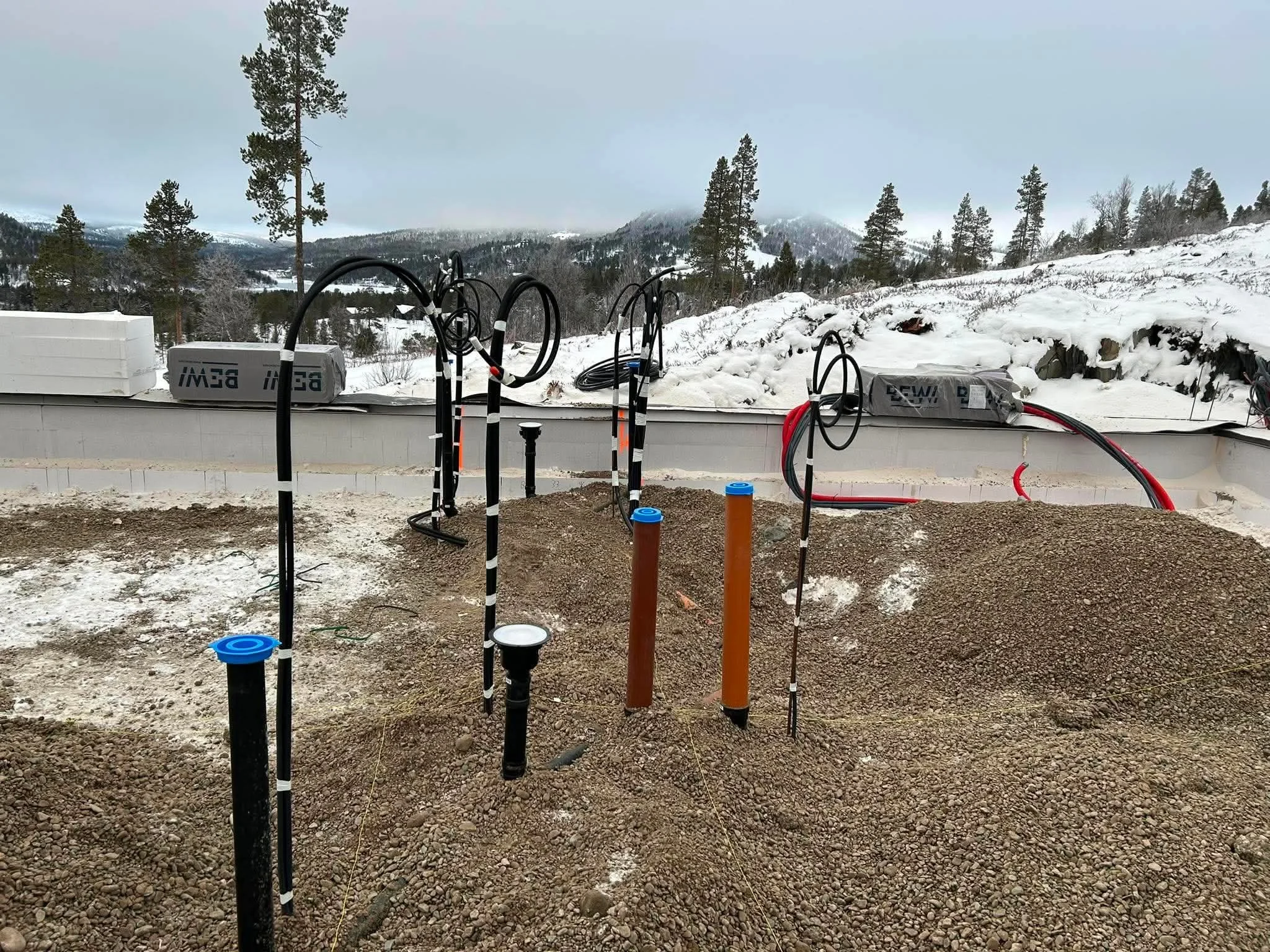 Grunnarbeid for et nybygg, med røropplegg til vann og kloakk, med snødekke rundt og fjell i bakgrunnen.
