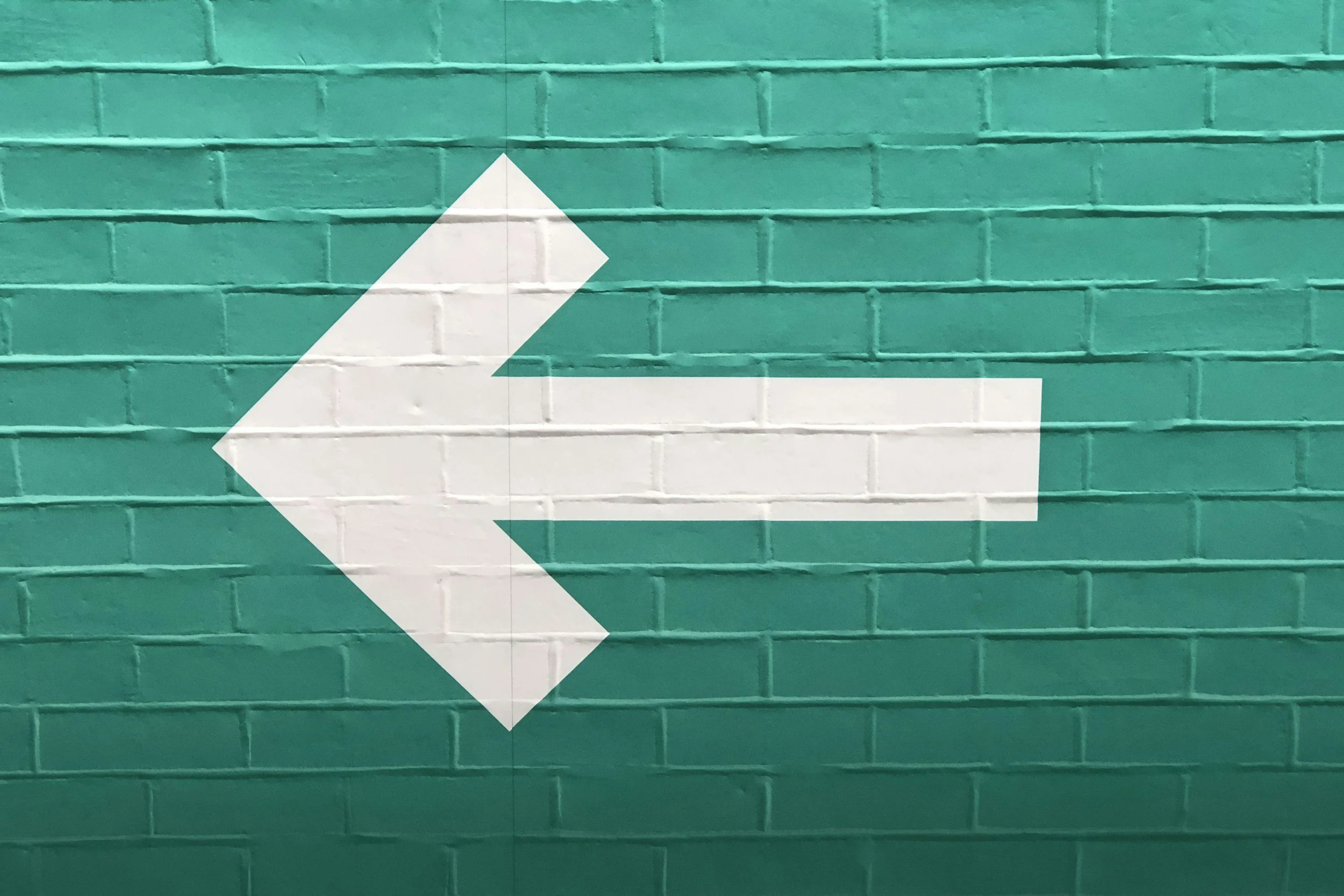 Flèche blanche pointant vers la gauche sur un mur en briques vert turquoise.