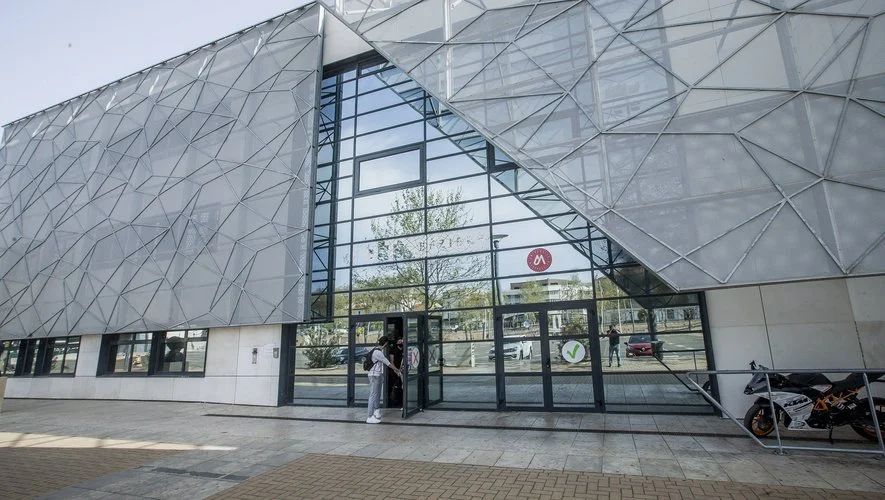Façade moderne d'un bâtiment avec une structure en métal et beaucoup de verre, avec une personne utilisant une borne d'information à l'entrée.