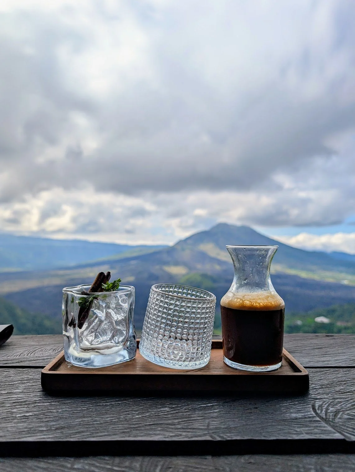 A wooden tray with a glass of dark coffee, a textured glass, and a small glass container with cinnamon sticks and mint, set on a wooden table with a scenic mountain and cloudy sky in the background.
