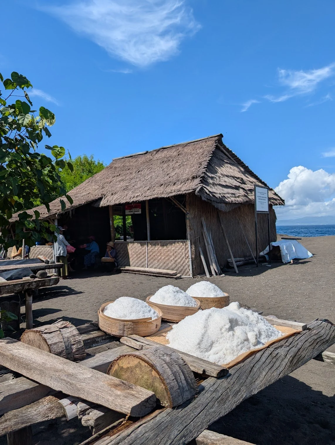 Traditional bamboo and thatch hut on a sandy beach near Kusamba Bali, with a blue sky, wooden trays with salt or white mineral salt, and people sitting nearby.