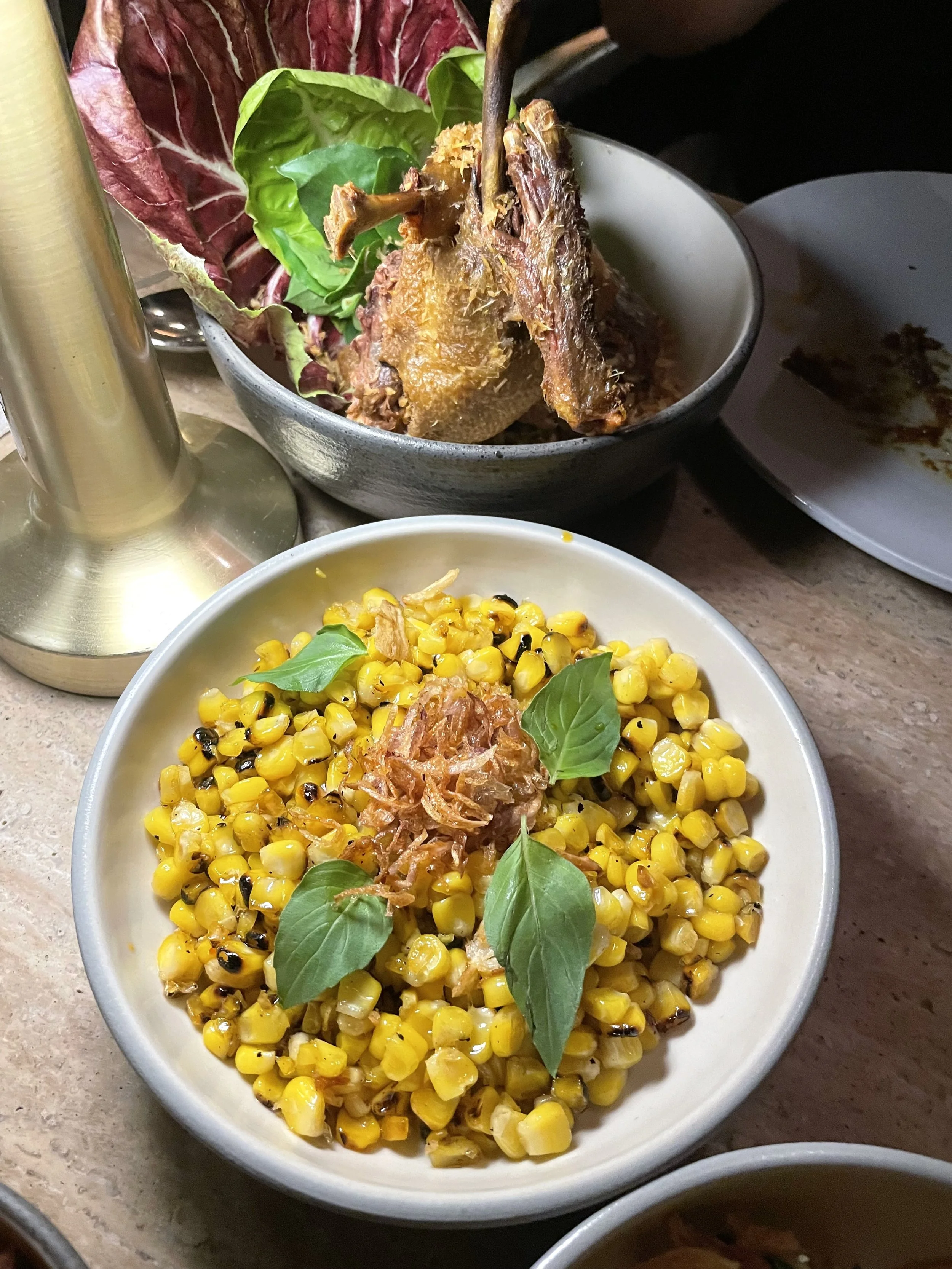 A bowl of grilled corn garnished with basil leaves and fried shallots, with a bowl of cooked meat and vegetables in the background.