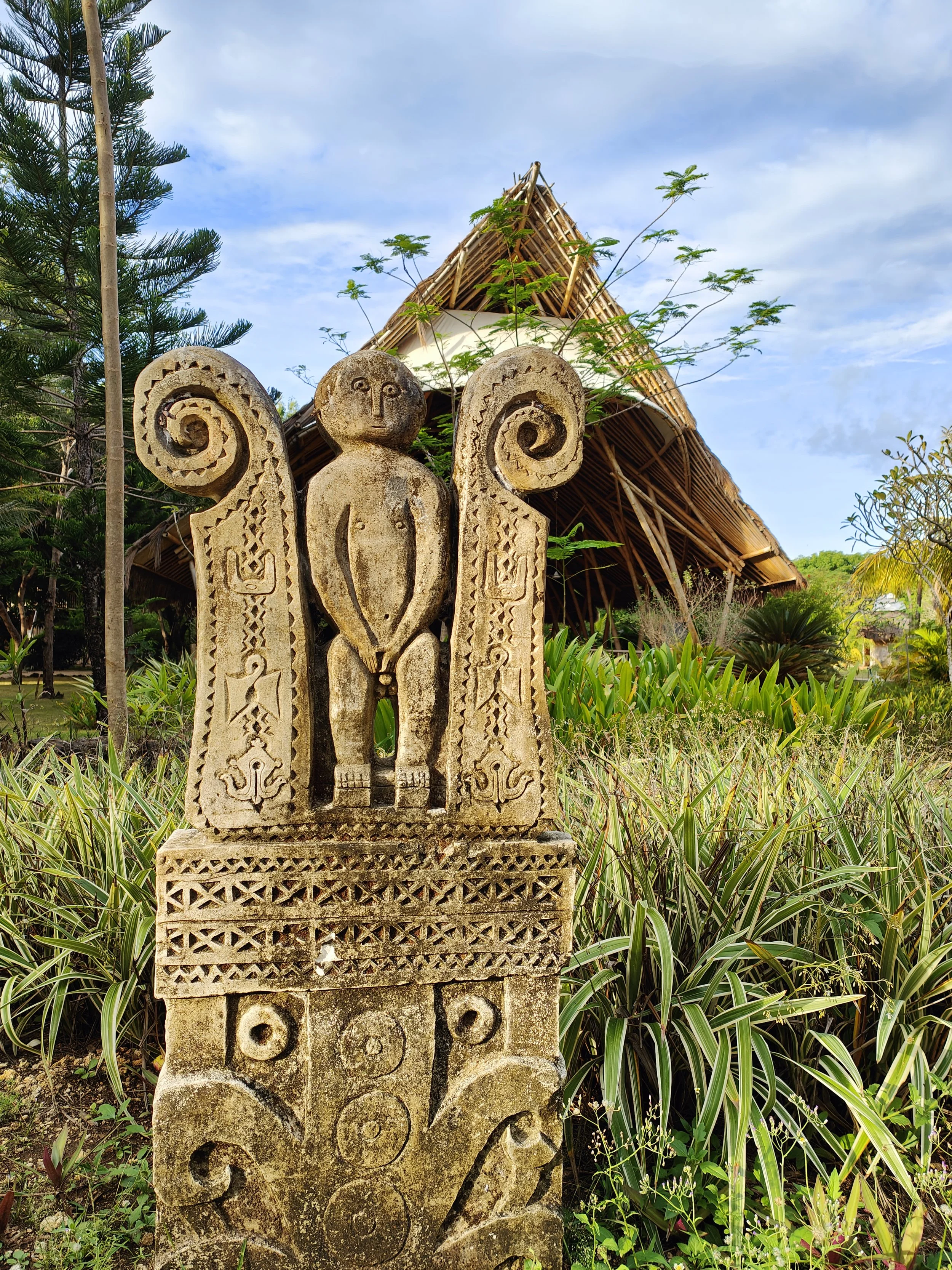 A stone sculpture of a human figure with large spiral ears, standing among lush green plants. In the background, there is a traditional Sumabense thatched roof structure and trees under a partly cloudy sky.