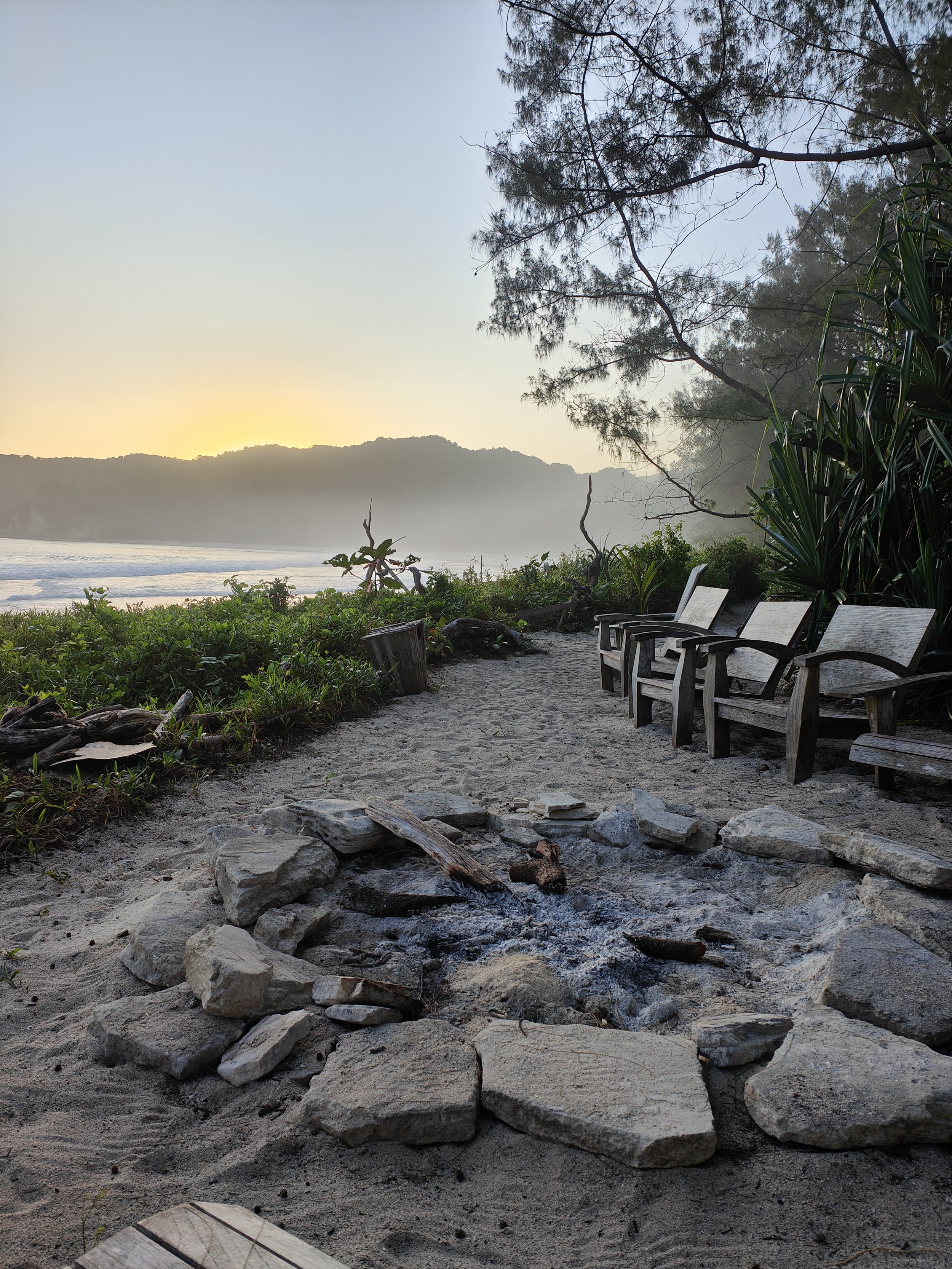 Tarimbang beach at sunset or sunrise with a fire pit made of stones in the foreground, surrounded by four wooden chairs, with trees and ocean waves in the background.