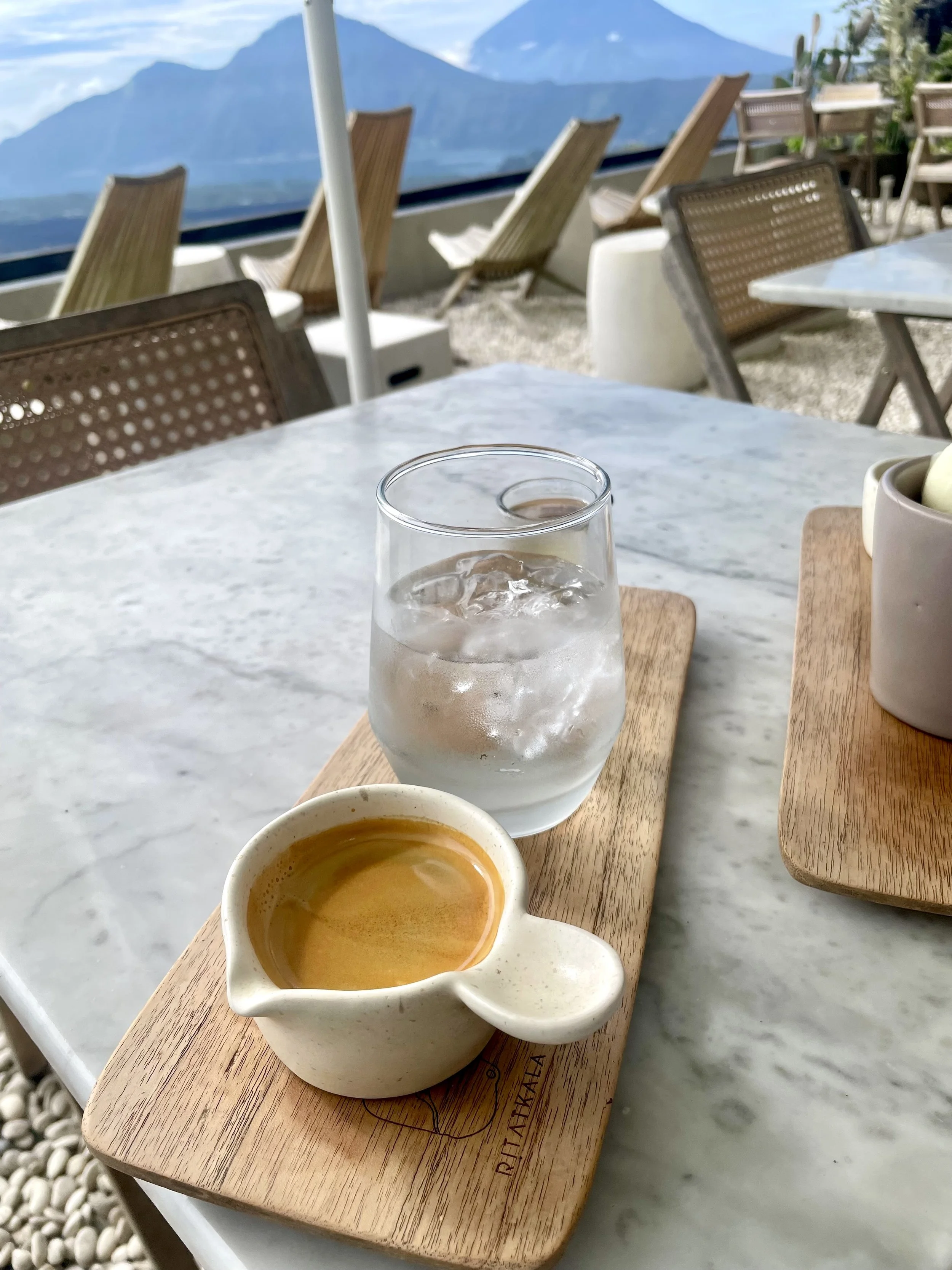 A marble table with a wooden tray holding a small cup of espresso and a glass of water with ice at an outdoor café, with Mt Batur , Bali the background.