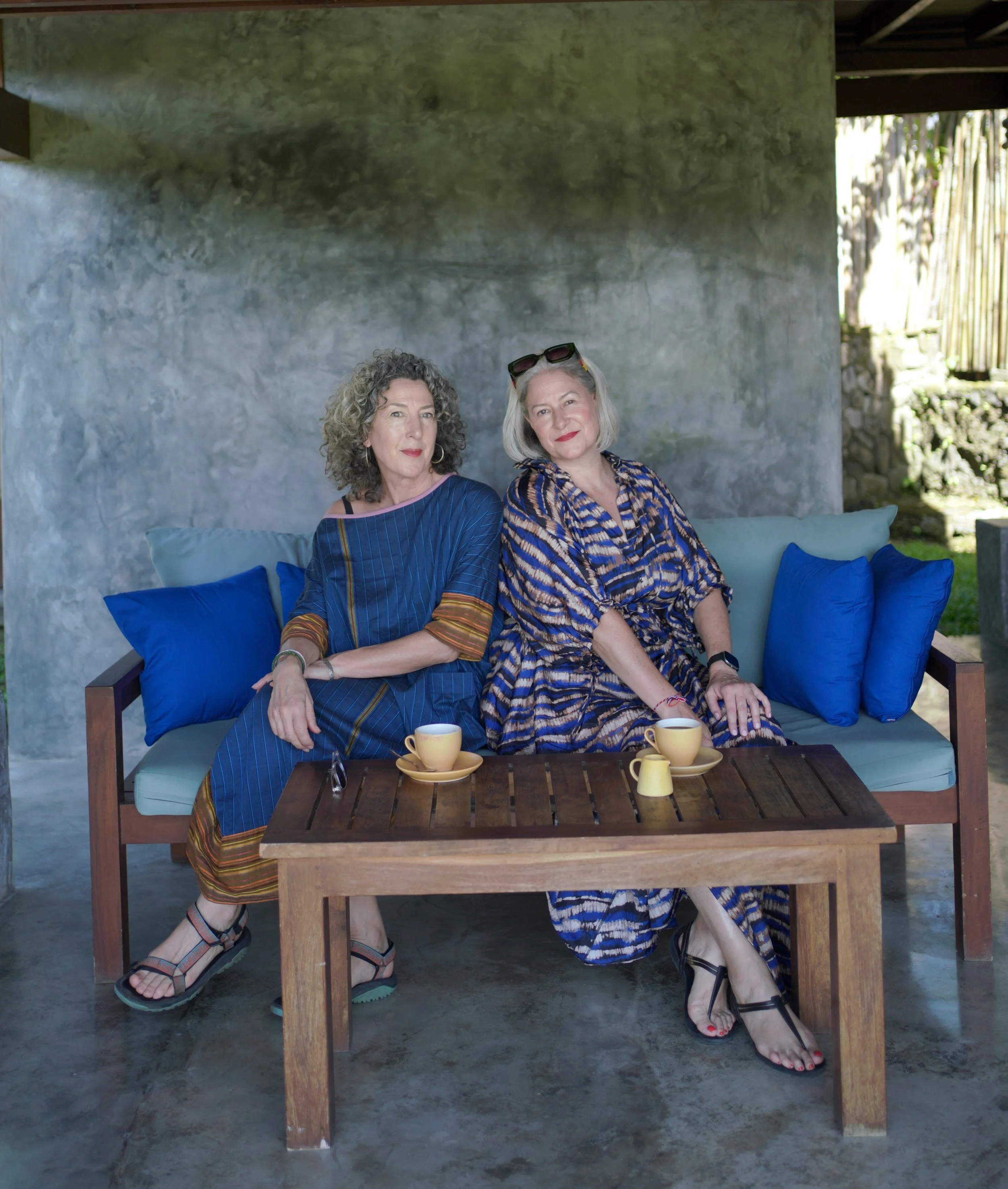 Two women sitting on a wooden bench with blue pillows, having coffee, under a shaded outdoor area with a stone wall behind them.
