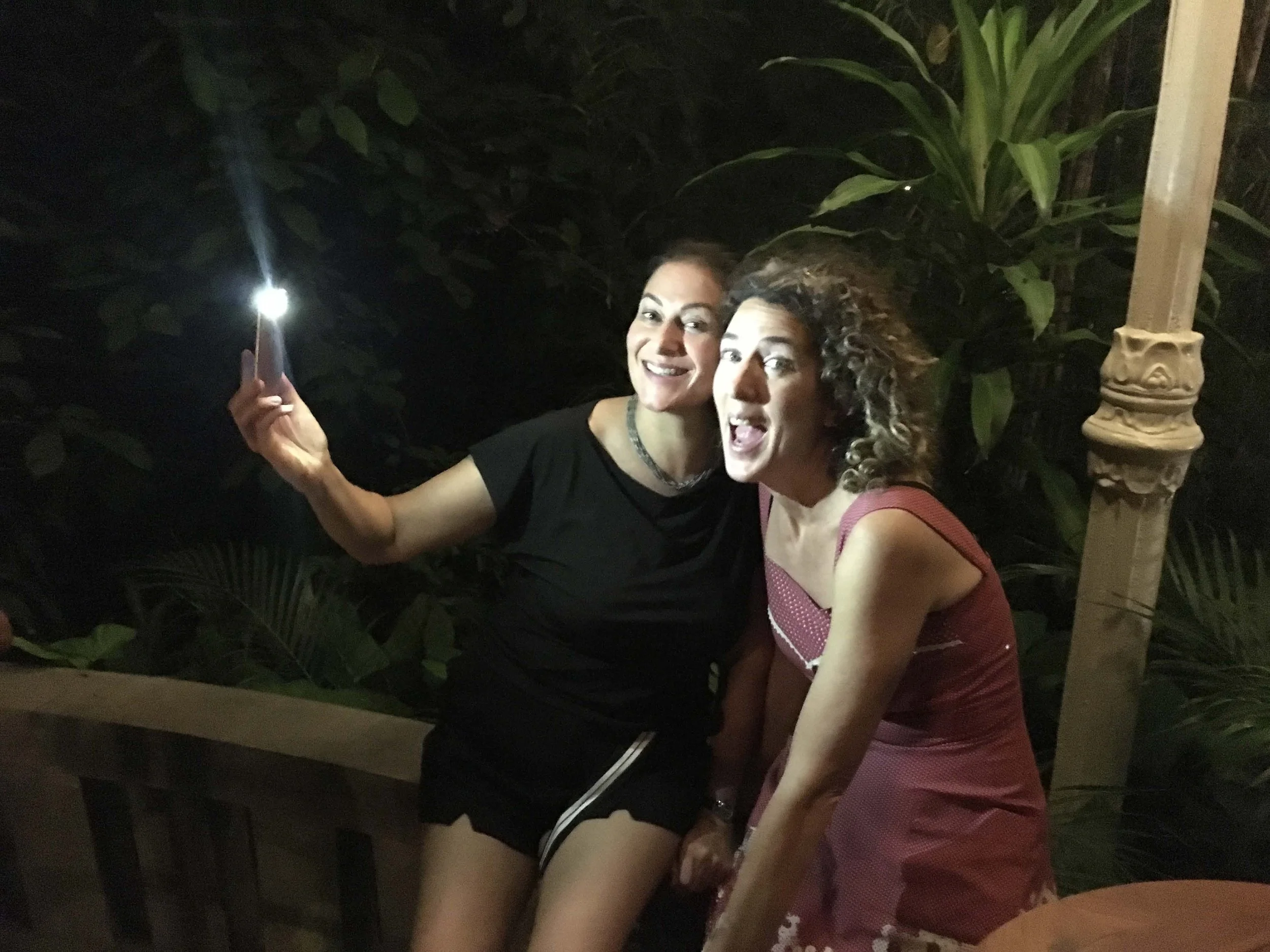 Two women smiling and taking a selfie at night outdoors, surrounded by jungle foliage, one holding a smartphone with flash on.