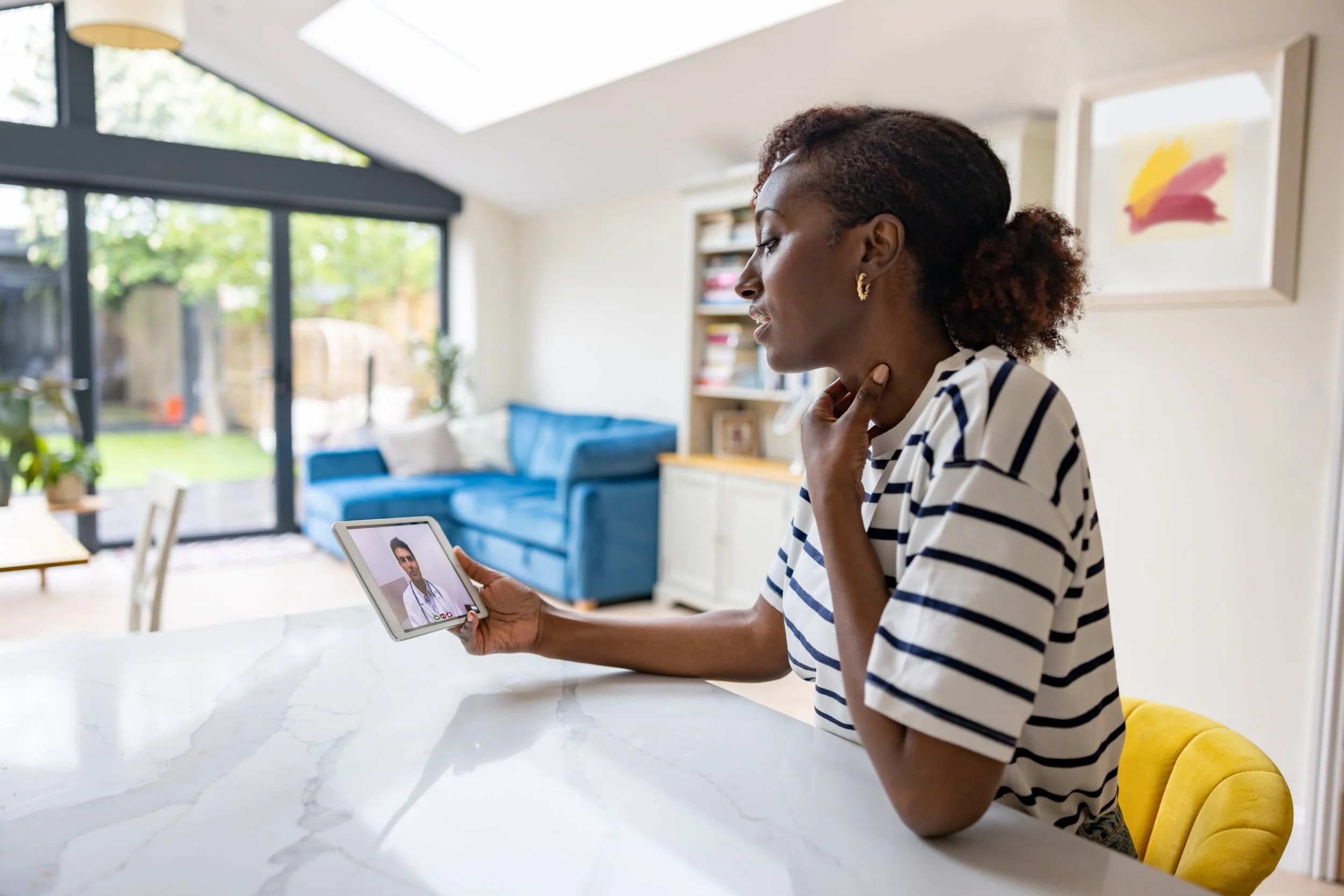 A woman in a striped shirt is holding a tablet with a video call to a doctor, in a bright kitchen with large windows, a blue couch, a colorful abstract painting, and plants.