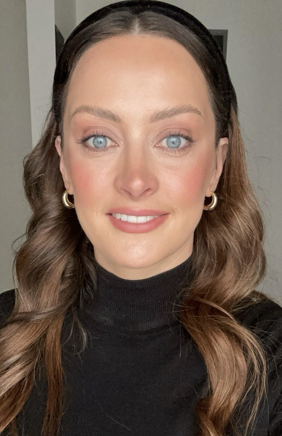 Close-up of a woman with long brown hair, blue eyes, wearing gold hoop earrings and a black turtleneck. She is smiling and looking directly at the camera.