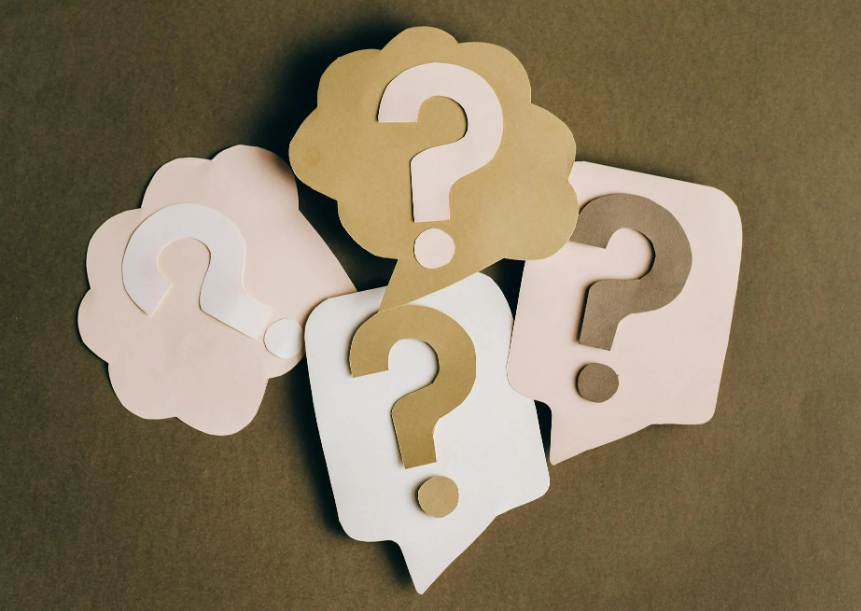 Plusieurs bulles de dialogue en papier avec des points d'interrogation, de couleurs blanche, beige et gris, disposées sur une surface brune.