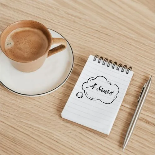 Une tasse de café sur une soucoupe, un carnet avec l'inscription "À venir" dans une bulle, et un stylo sur une surface en bois.