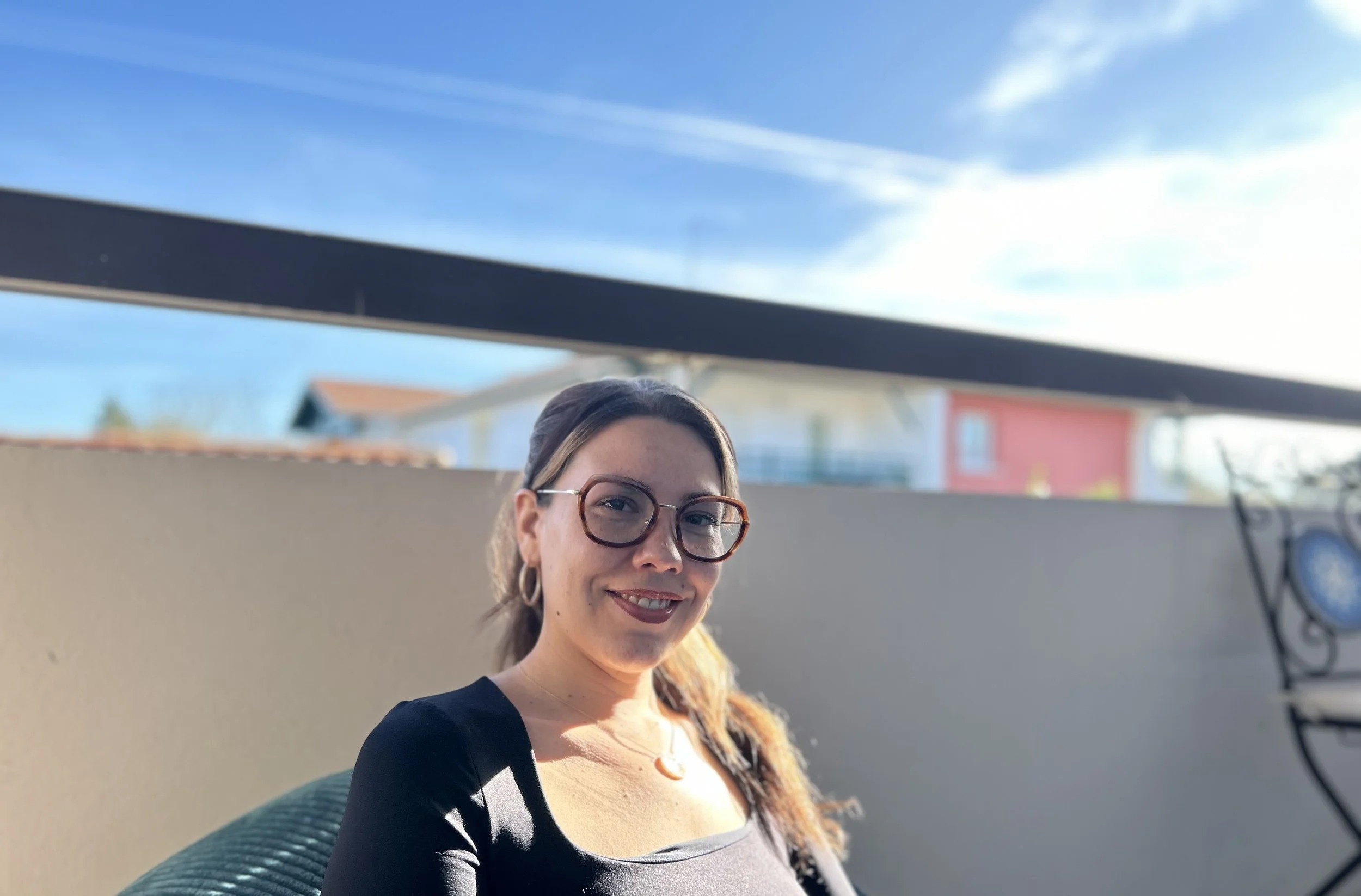 Une femme souriante portant des lunettes, assise à l'extérieur sous un ciel bleu avec quelques nuages, avec un mur blanc derrière elle et une chaise en métal à droite.