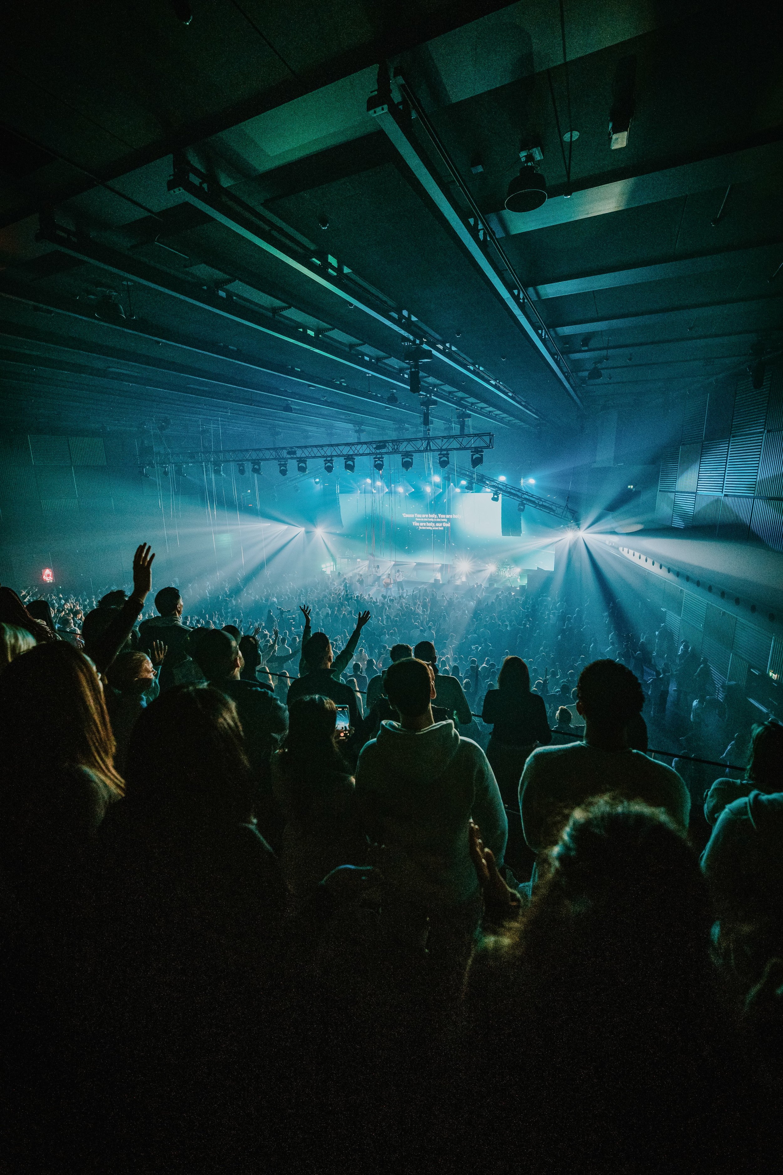 Menschenmenge bei einem Konzert in einem dunklen Saal mit Scheinwerfern, Lichtern und Bühne im Hintergrund.