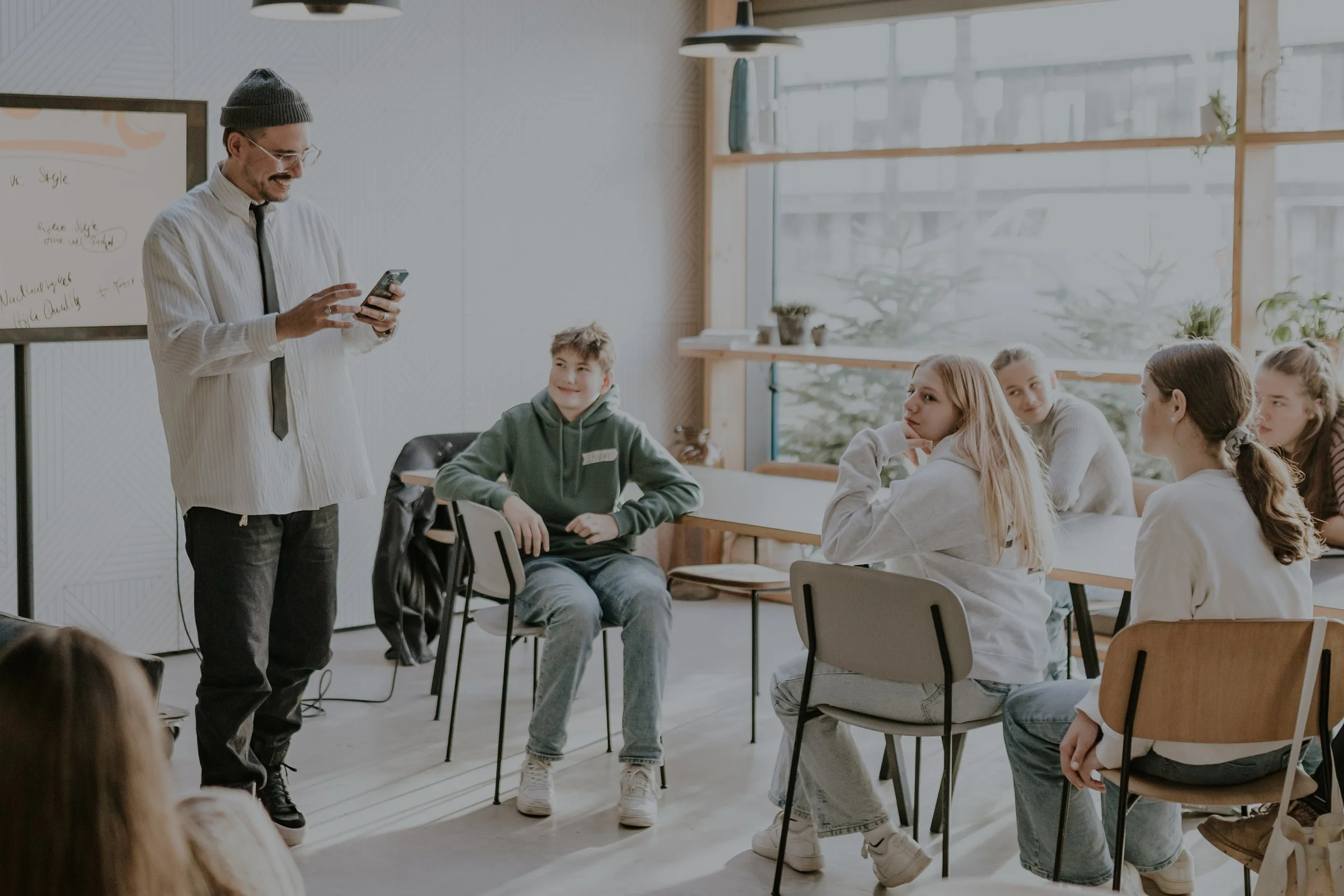 Ein Mann in einem Klassenraum zeigt auf seinem Smartphone, während fünf Jugendliche ihm zuhören. Der Raum hat große Fenster und eine Tafel im Hintergrund.